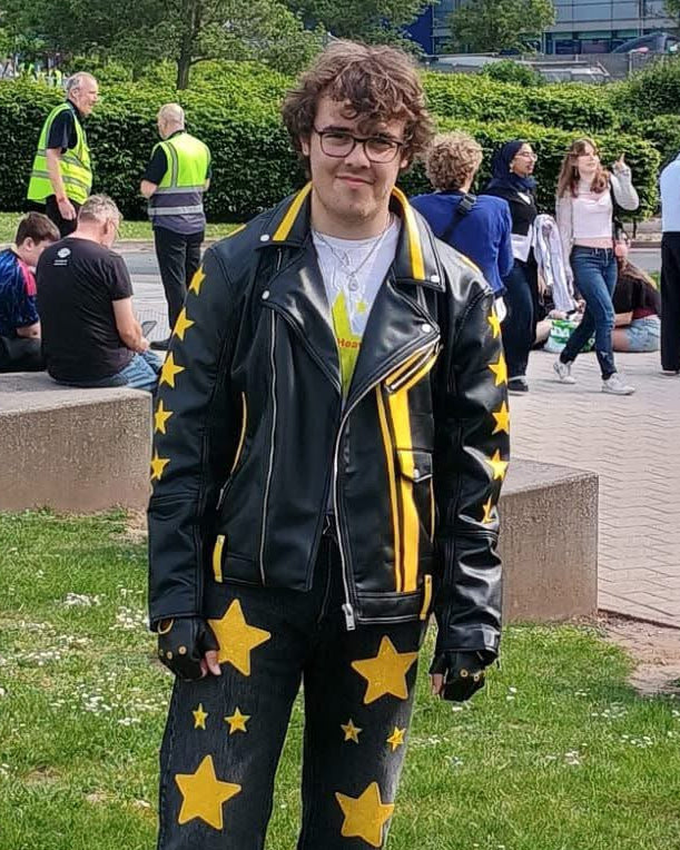 Person wearing a black outfit with yellow star patterns standing in a park-like setting with trees and a building in the background.