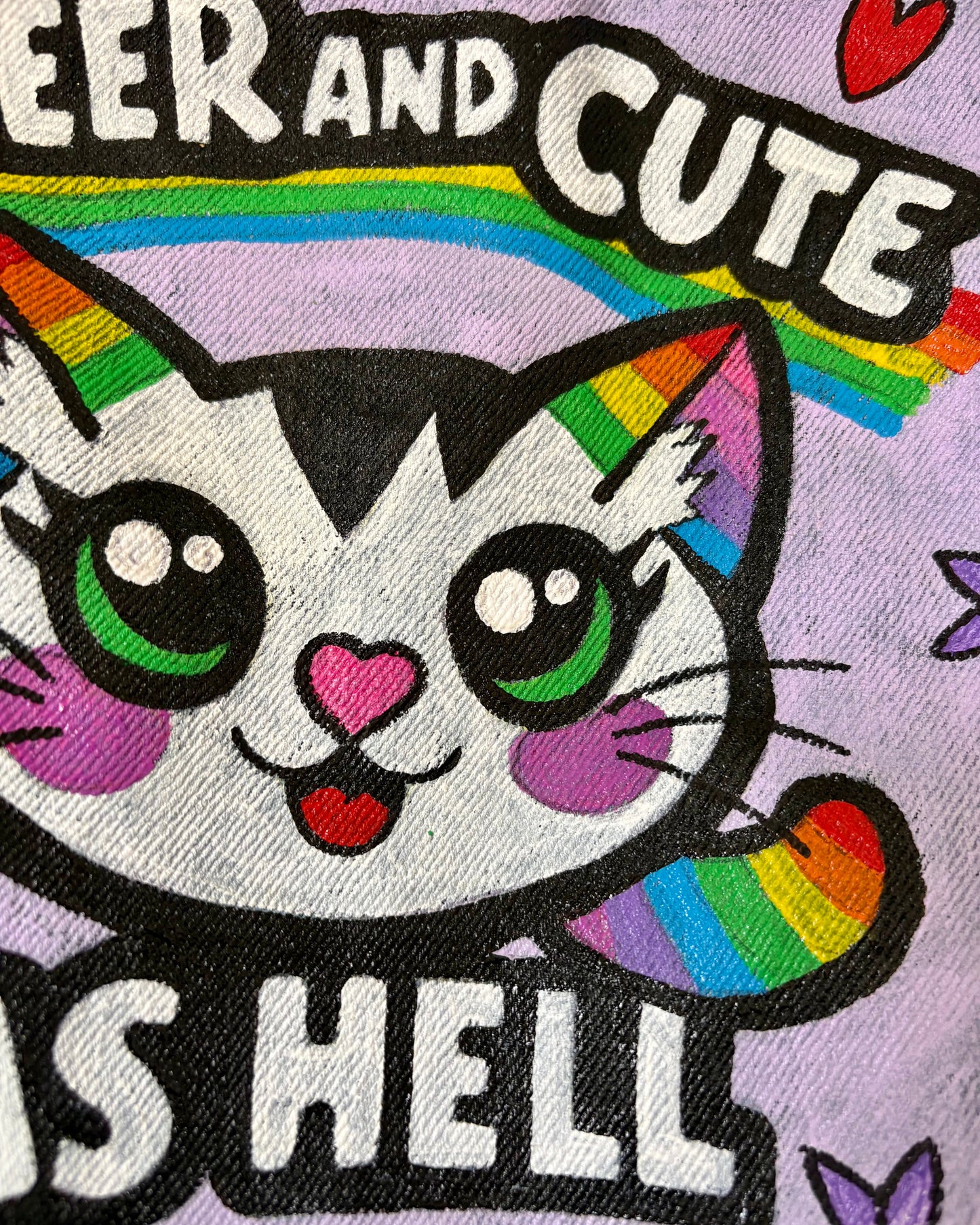 Close up view of a pastel purple panel on a hand-painted upcycled denim jacket featuring a rainbow cat, hearts, flowers, and the words “Queer and Cute as Hell”