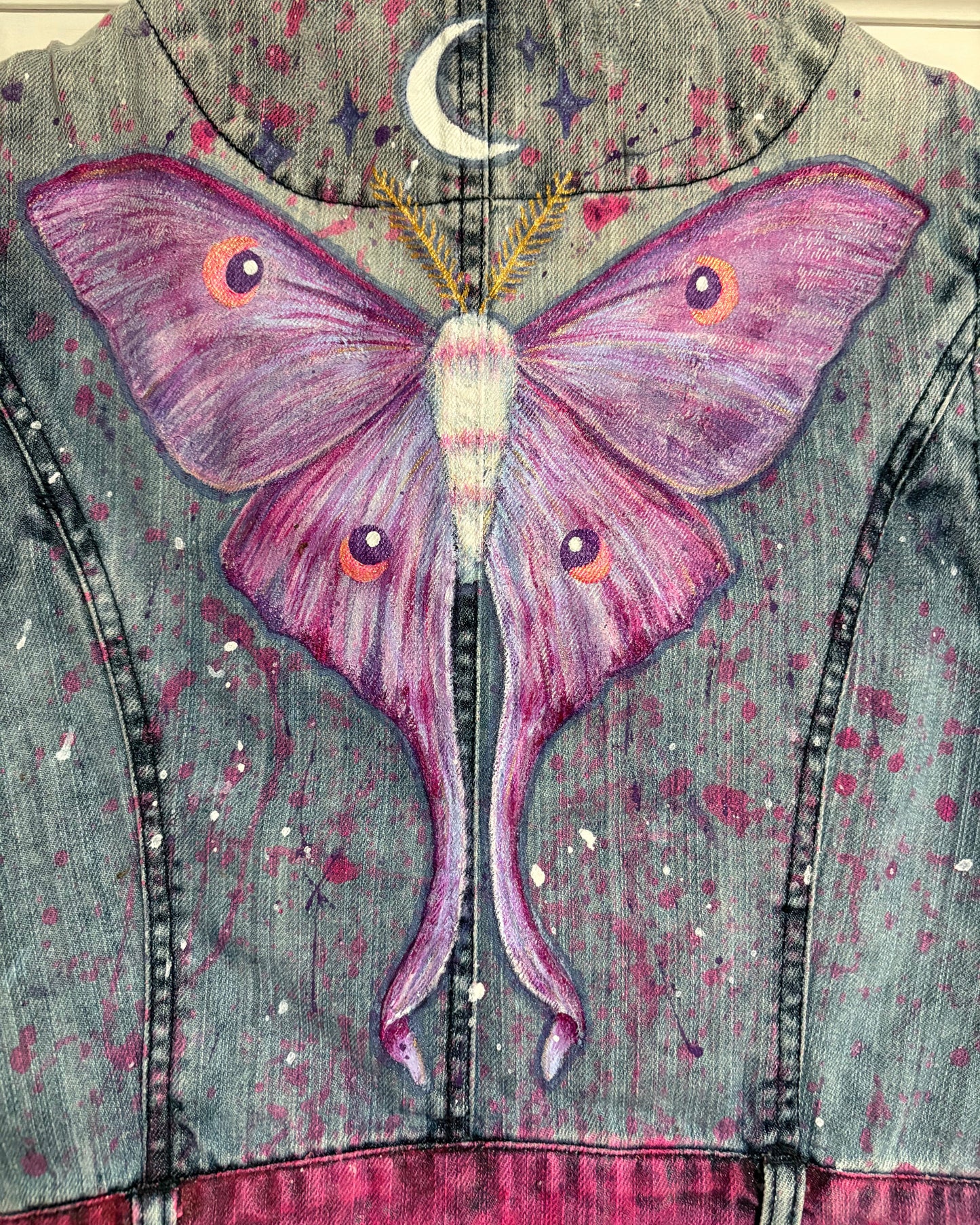 A close-up of the back of a denim jacket featuring a large painted purple and pink moon moth, with a crescent moon and stars on a distressed gray-blue denim background with pink paint splatter details.