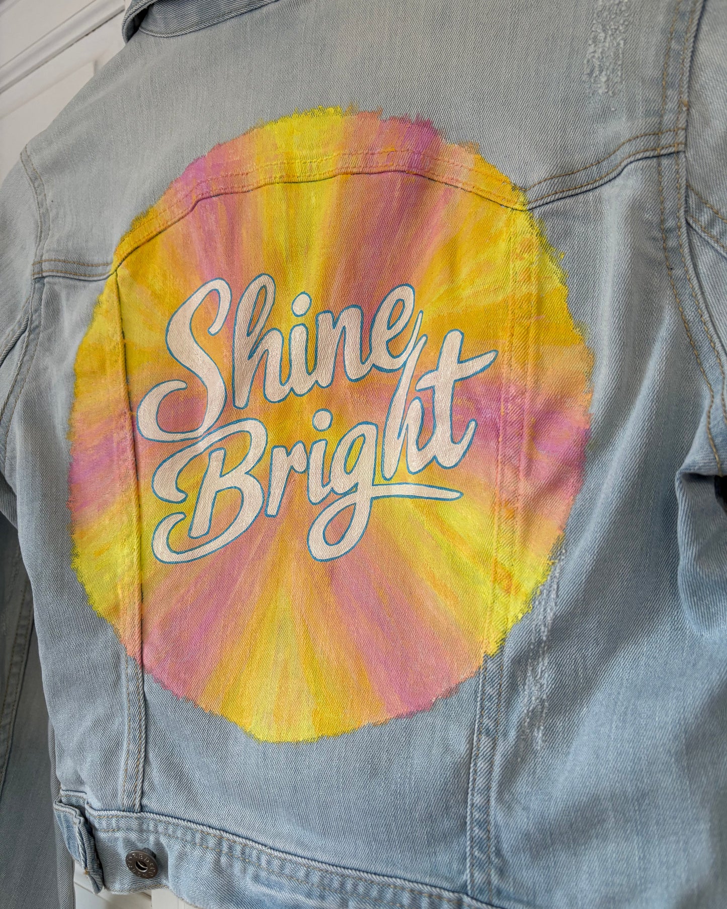 Close up of denim jacket with a colourful hand painted sunburst and 'Shine Bright' written in the centre.