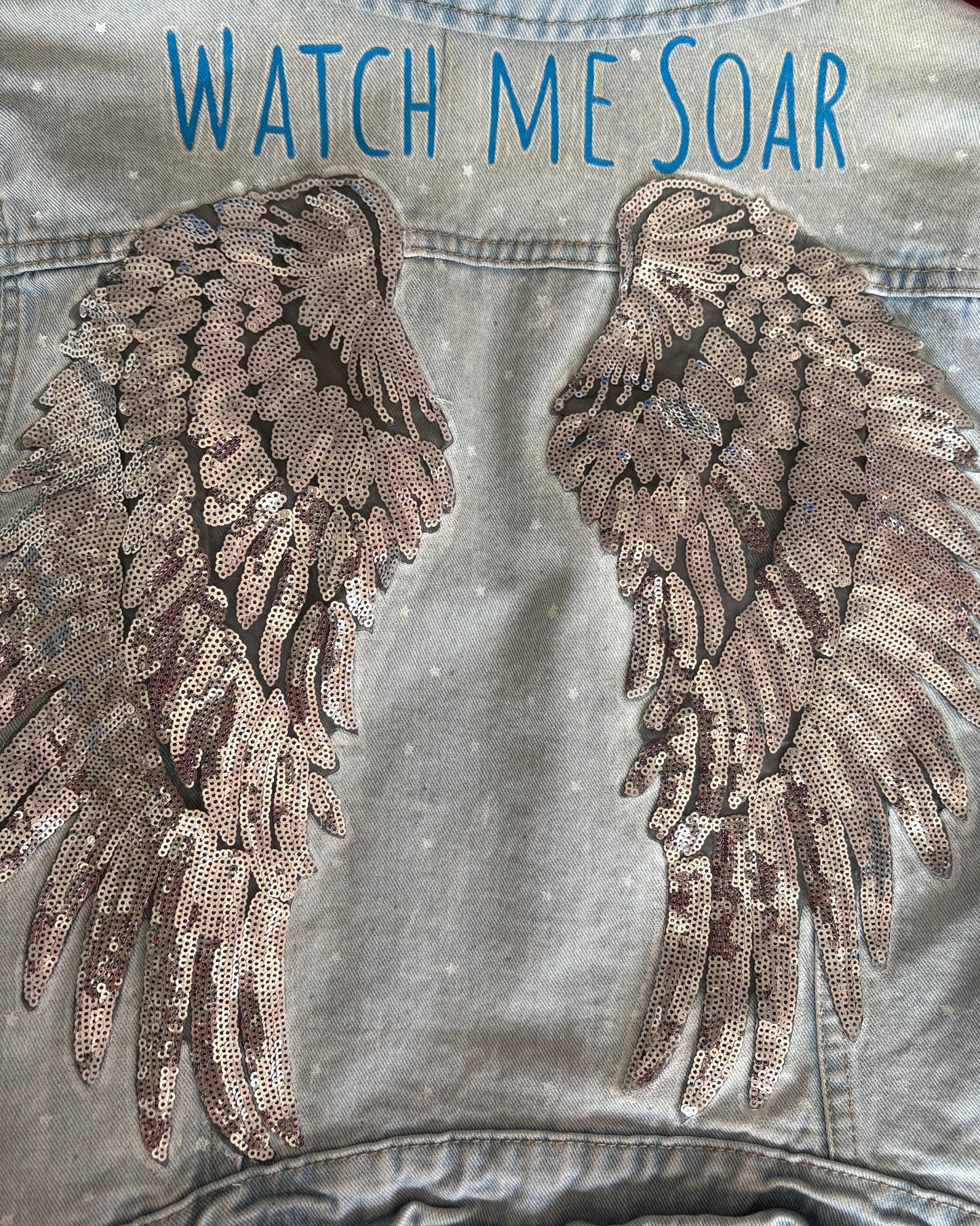 Denim jacket with sequin wings and 'Watch Me Soar' text painted on.