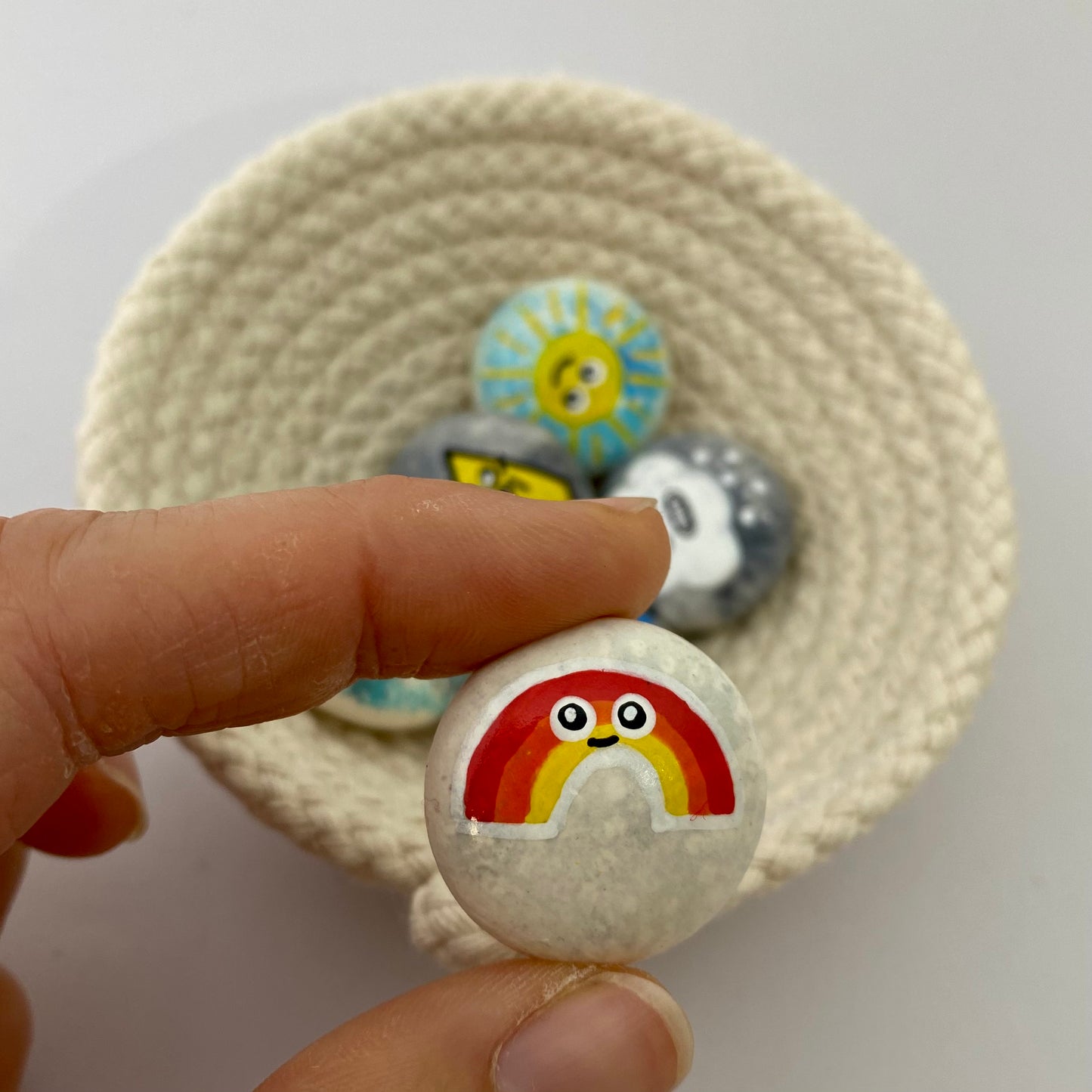 6 small hand painted weather themed pebbles in a rope bowl, the Rainbow pebble is being held close to the camera