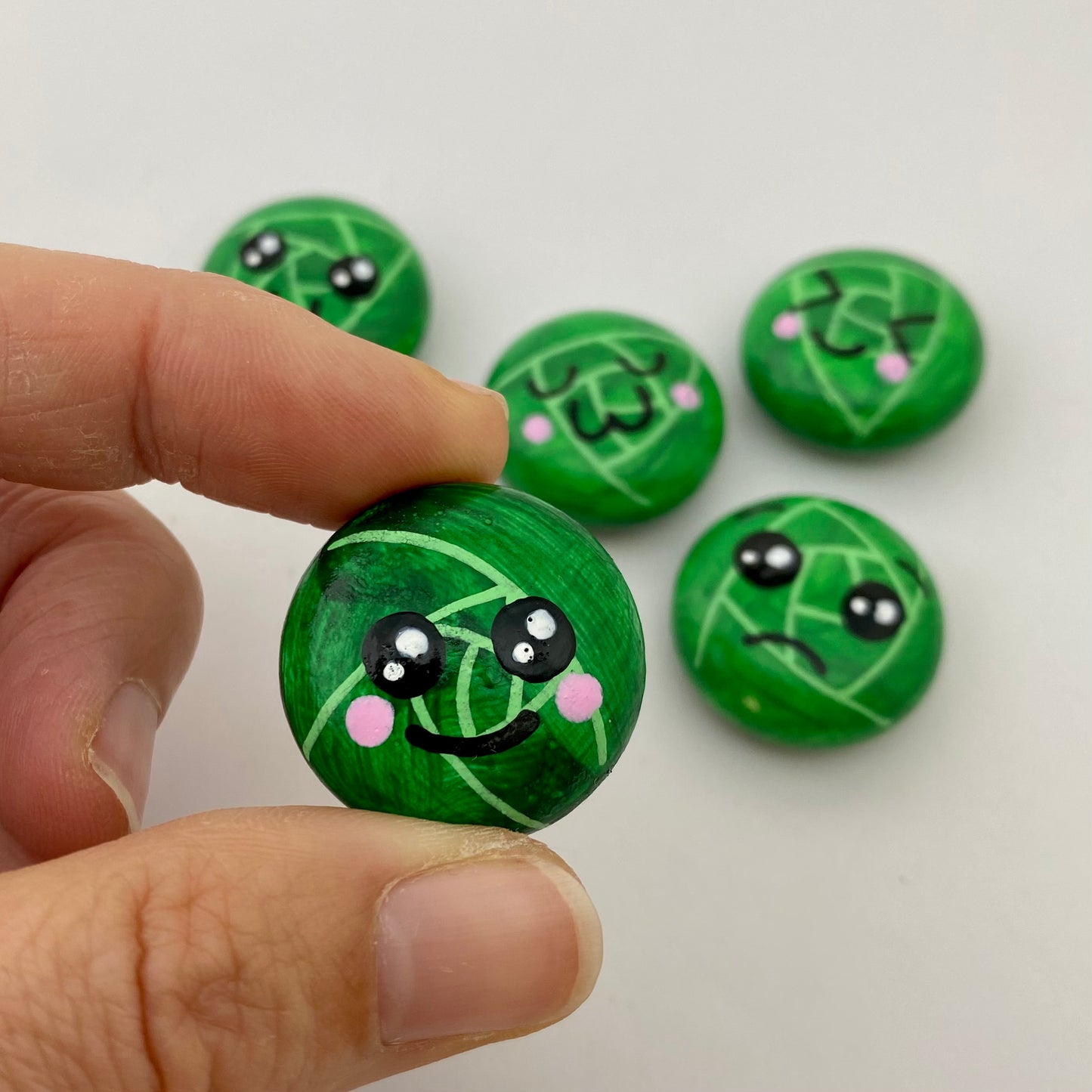 5 Hand painted tiny sprout pebbles each with a different cute faced expression. One is being held closer to the camera, this one has a smily face