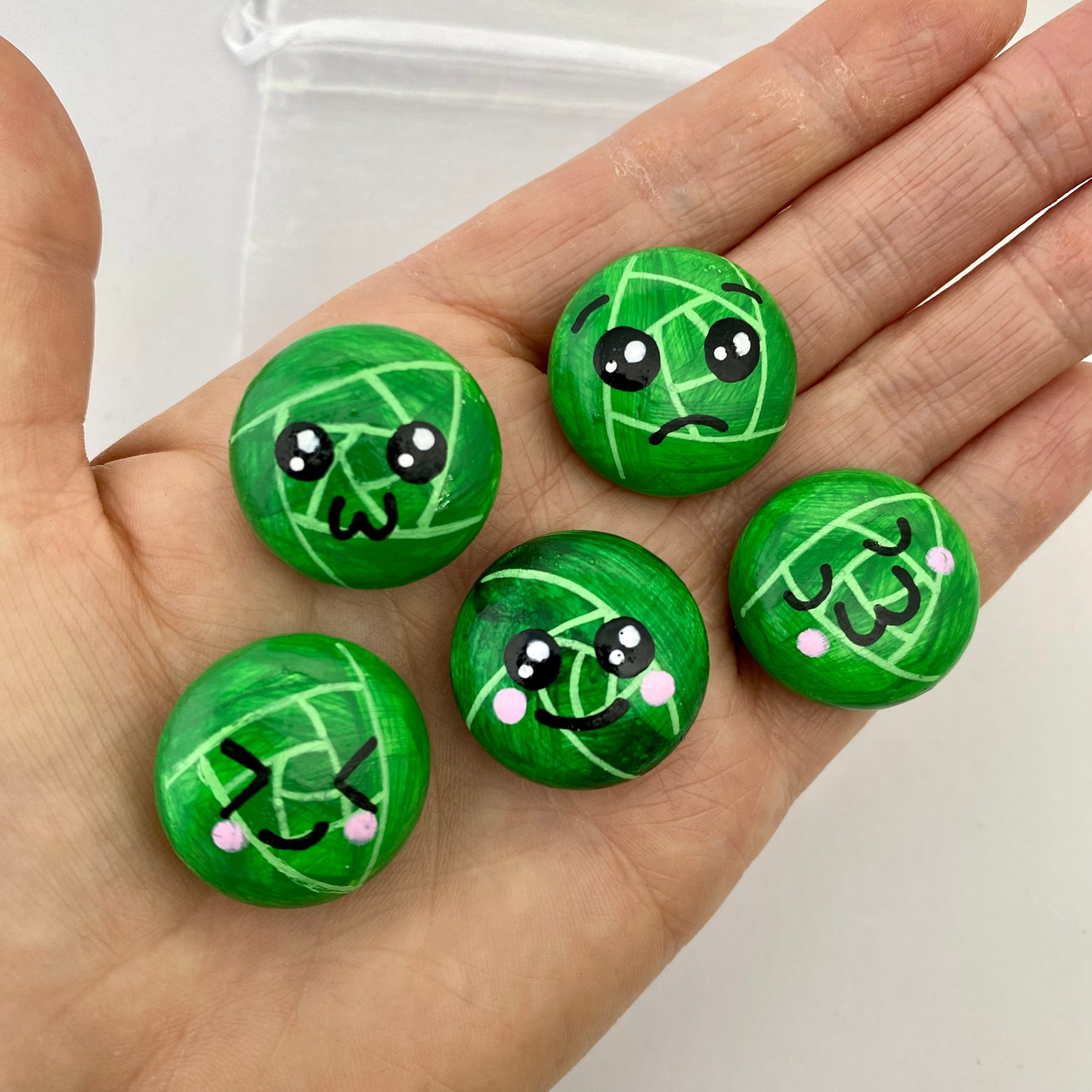 5 Hand painted tiny sprout pebbles each with a different cute faced expression held in a hand