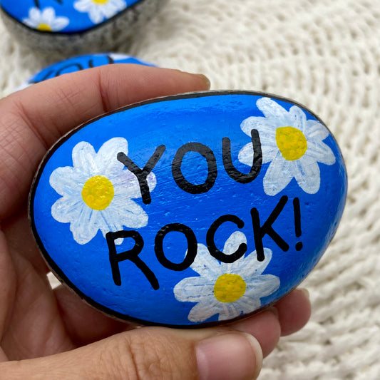 A rock painted blue with daisies and the words "You Rock!" painted over the top.