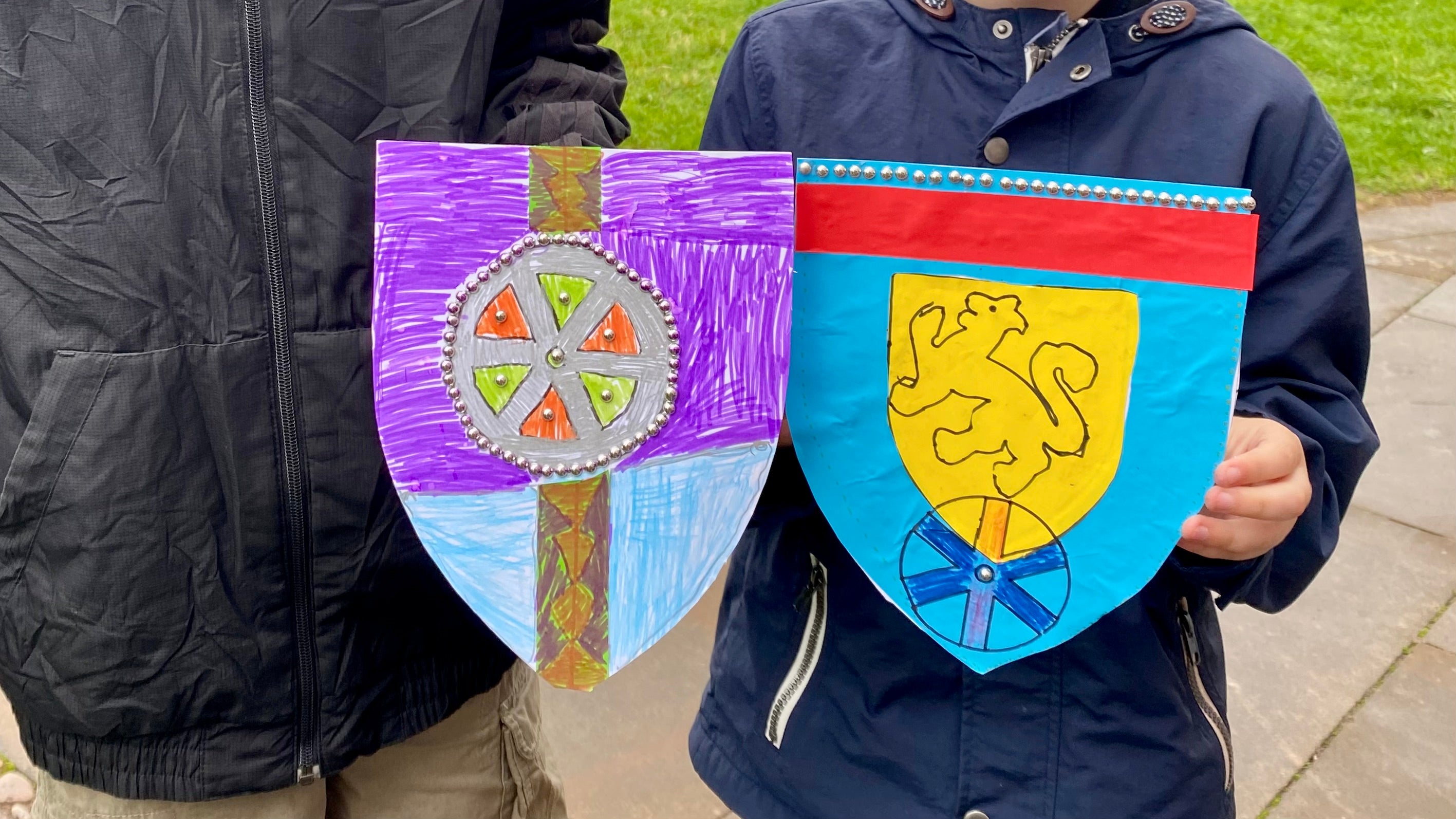 Two cardboard shields being held by children. One shield is purple and blue with a brown stripe and a wheel motif in the centre with silver stud accents. The other shield is blue with a red stripe across the top and a line of silver studs along the top, in the centre is a smaller yellow shield shape with a lion motif drawn in.
