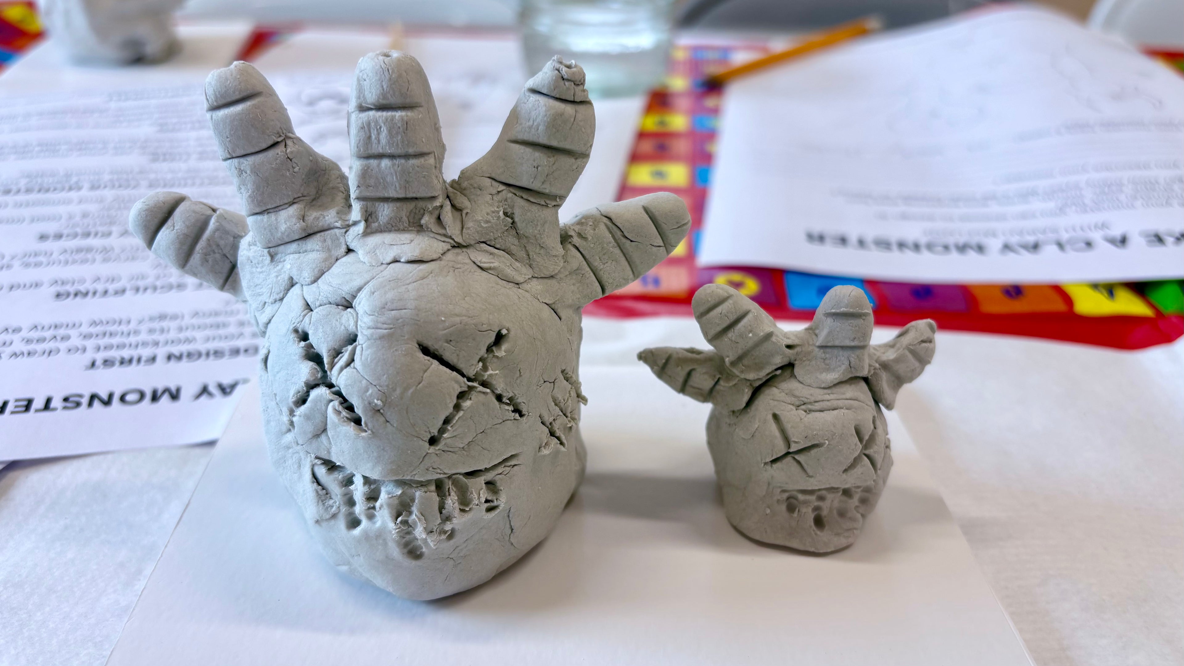 Two clay models of monsters on a table with how to worksheets in the background.