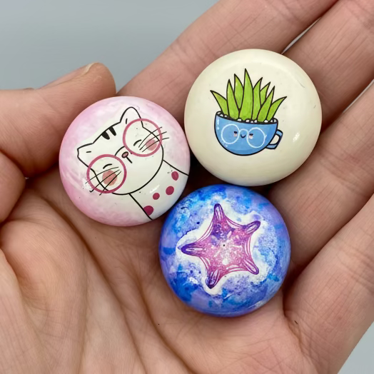 A hand holding 3 colourful mini pebbles with various designs on. A cartoon cat with pink glasses, a succulent in a teacup, a starfish.