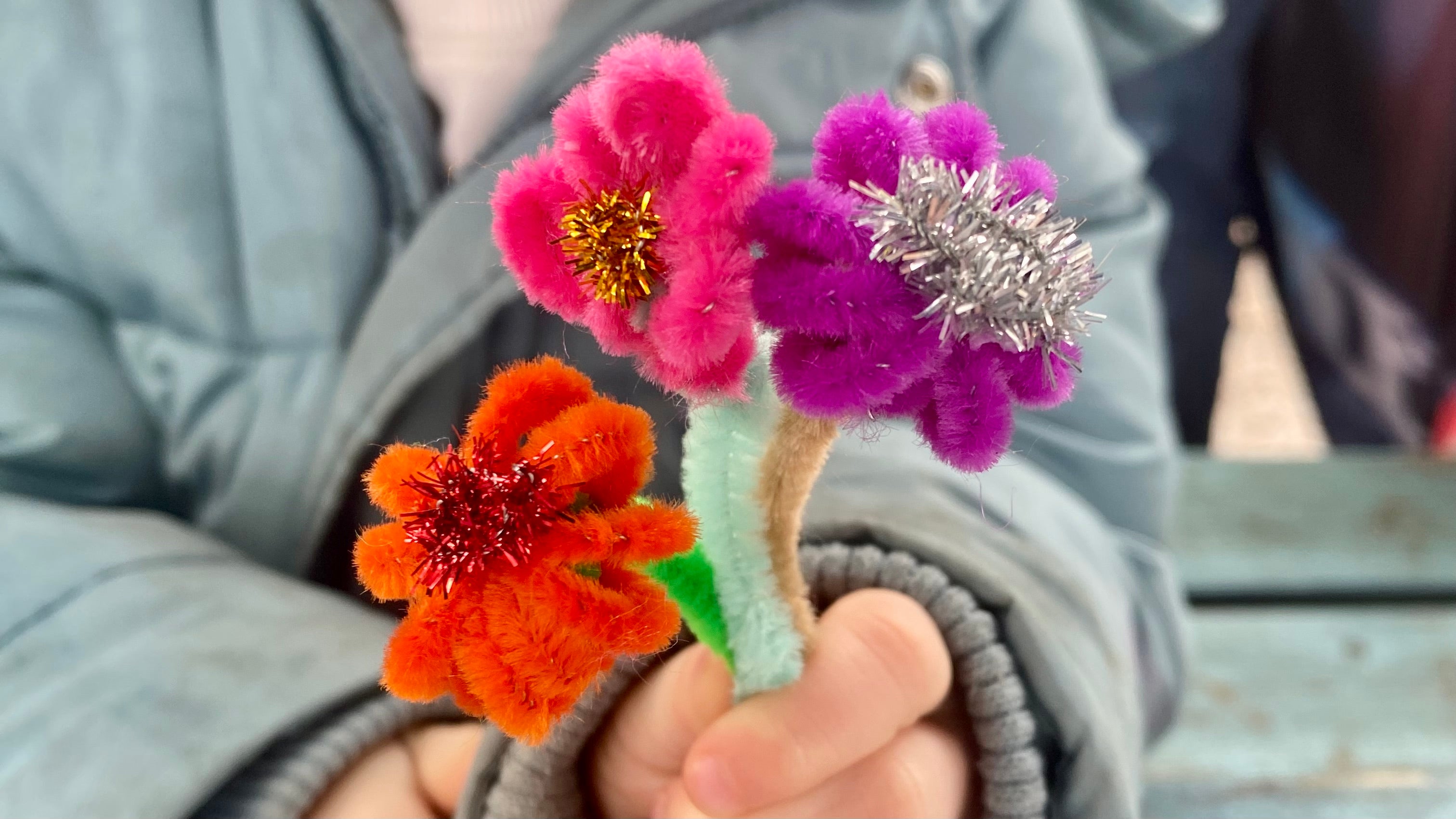 A child's hand holding 3 small flowers made from pipecleaners, the centre of each flower is made from a metallic sparkly pip cleaner, the flowers are red, pink and purple and the stems are different shades of green and brown.