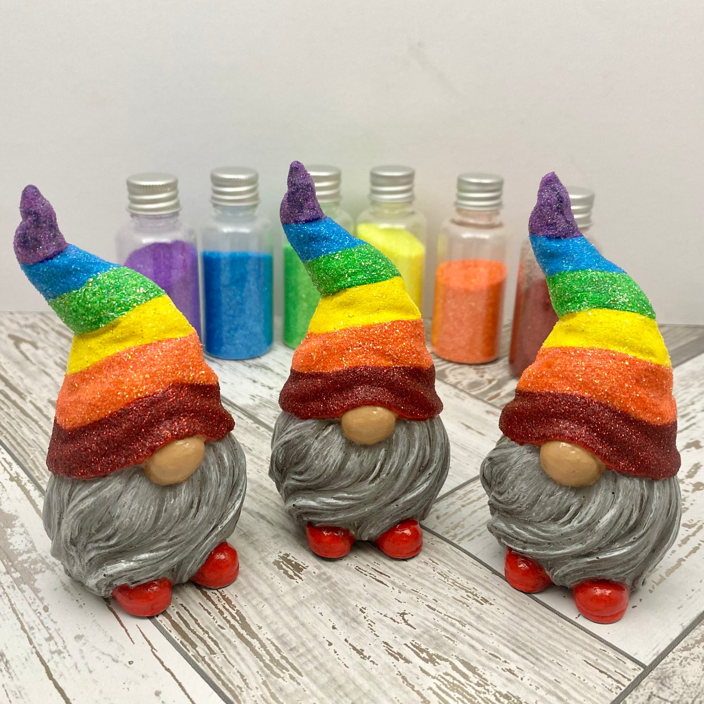 3 Small hand painted gonk statues with Glittery Rainbow hats, grey beards and red shoes. Behind them stands 6 different coloured bottles of glitter. 