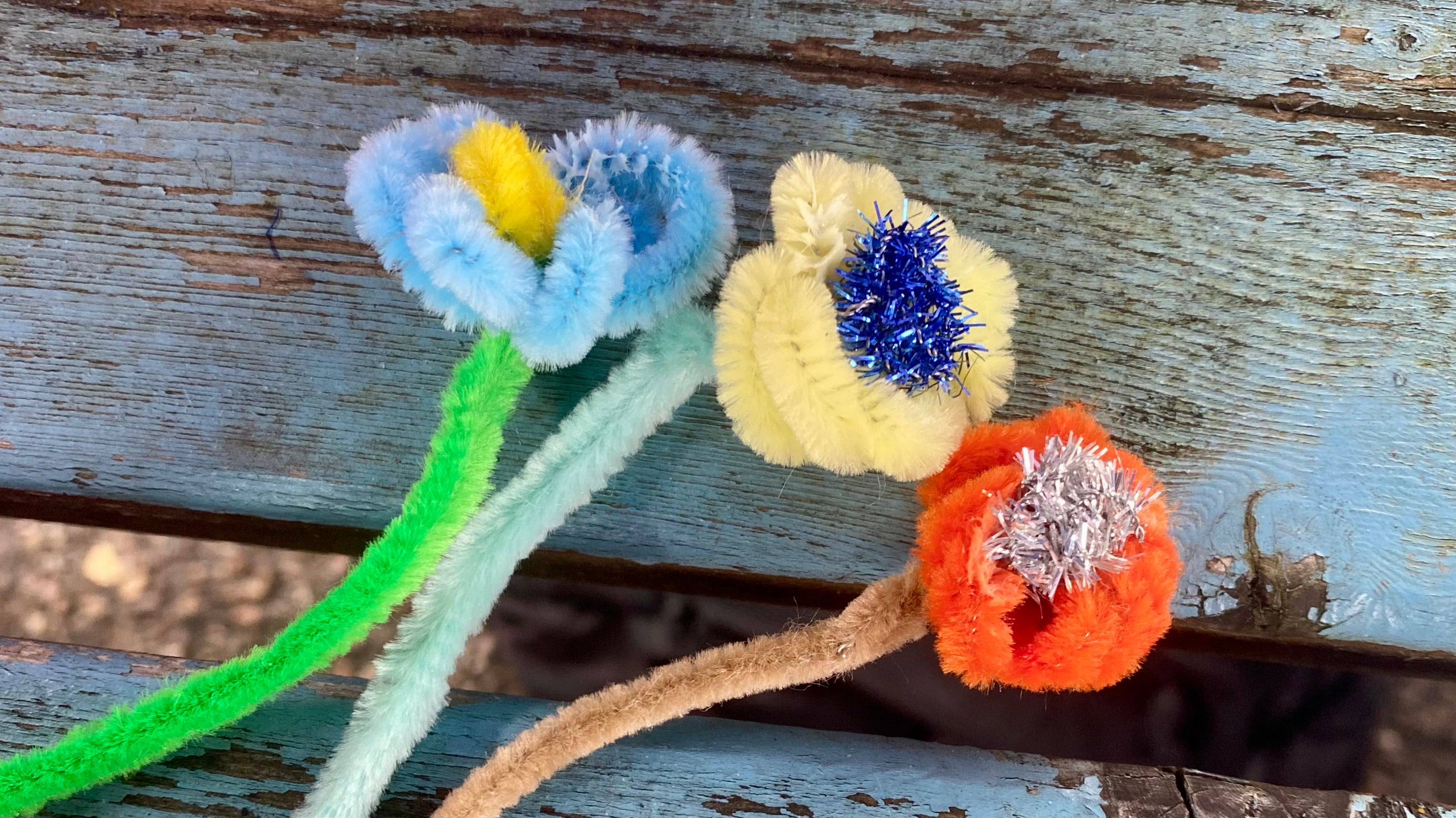 3 small flowers made from pipecleaners laying on a woooden bench, the centre of each flower is made from a metallic sparkly pipe cleaner, the flowers are blue, yellow and orange and the stems are different shades of green and brown.