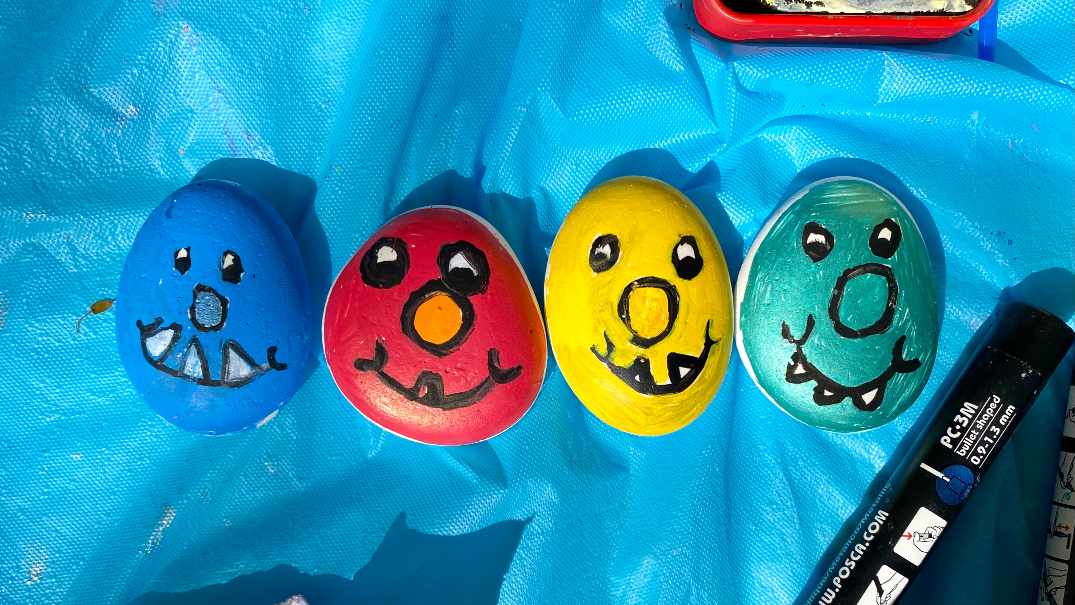 4 painted pebble monsters, one blue, one red, one yellow and one green. On a tablecloth with paints and pens.