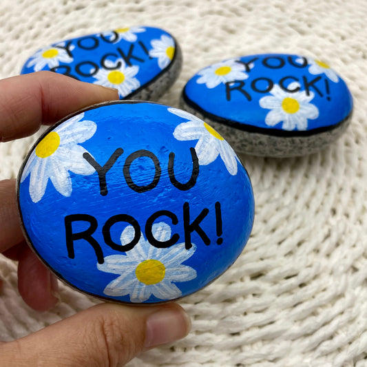 3 rocks painted blue with daisies and the words "You Rock!" painted over the top.
