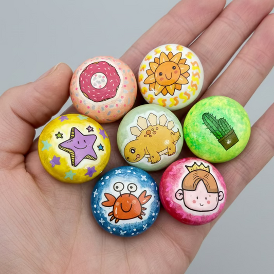 A hand holding 7 colourful mini pebbles with various designs on. A doughnut, a sunshine, a star, a dinosaur, a cactus, a crab and a princess.