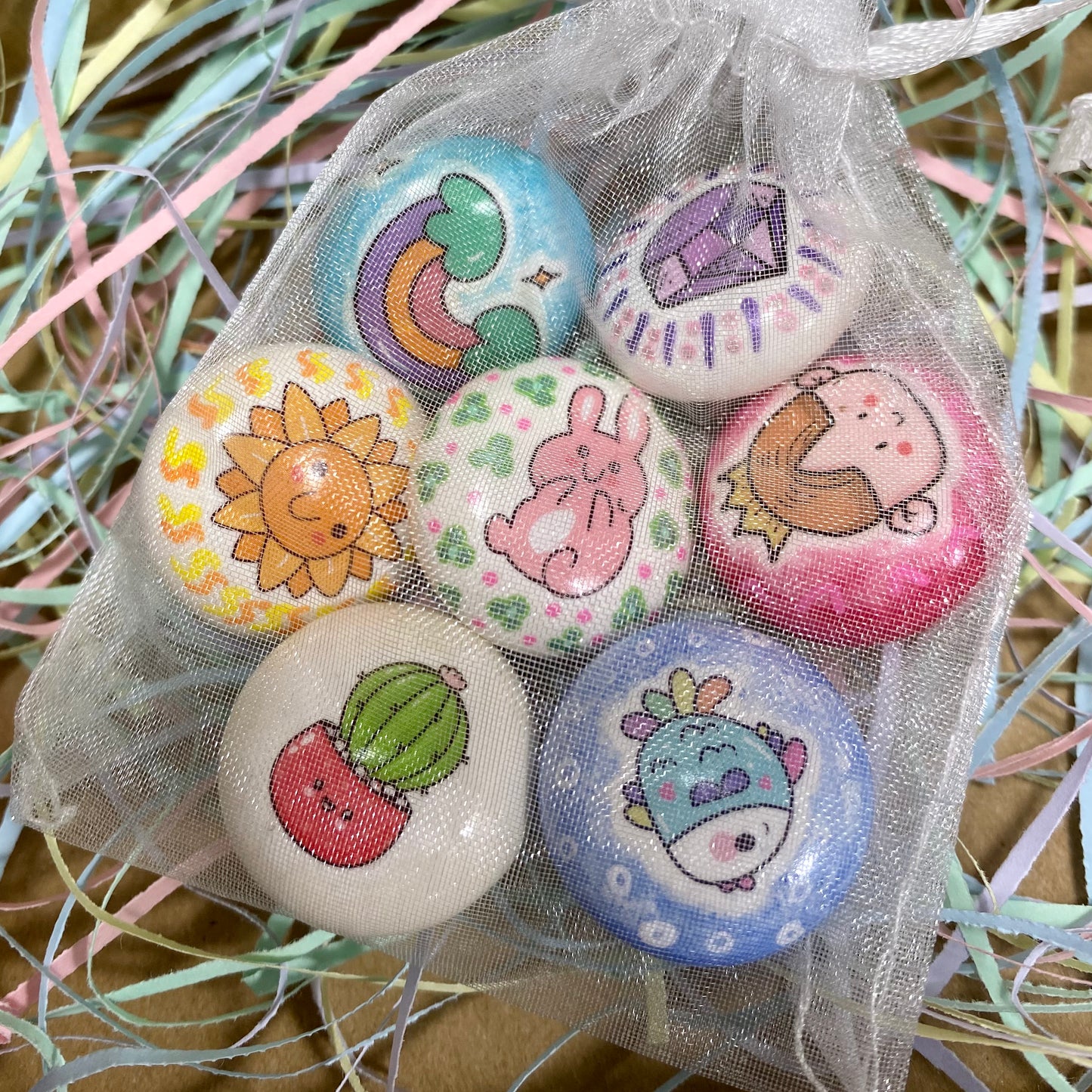 An organza bag surrounded by colourful shredded tissue paper. Inside the bag are 7 colourful mini pebbles with various designs on. A rainbow, a purple gem, a sunshine, a pink cartoon bunny, a princess, a potted cactus and a rainbow fish.
