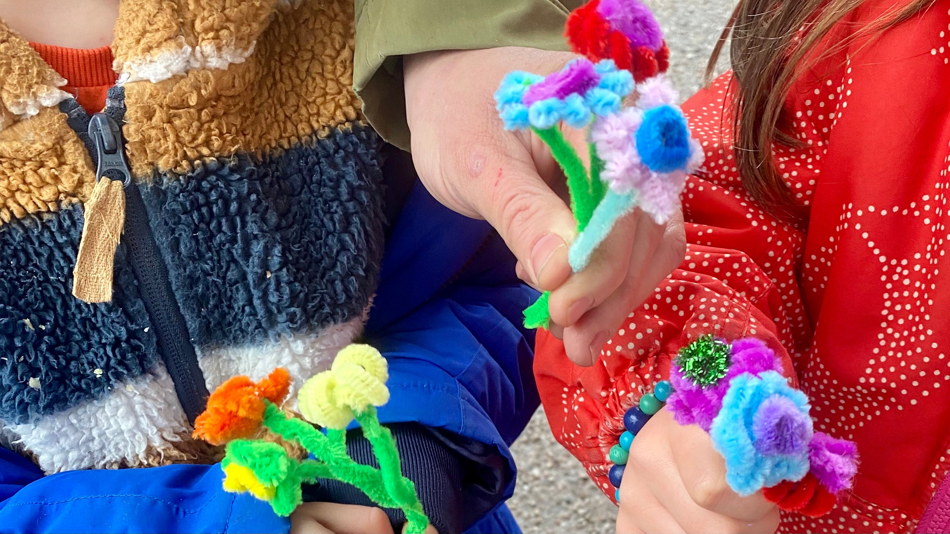 3 children's hands each holding 3 small flowers made from pipecleaners, each flower is made from 3 different coloured pipecleaners.