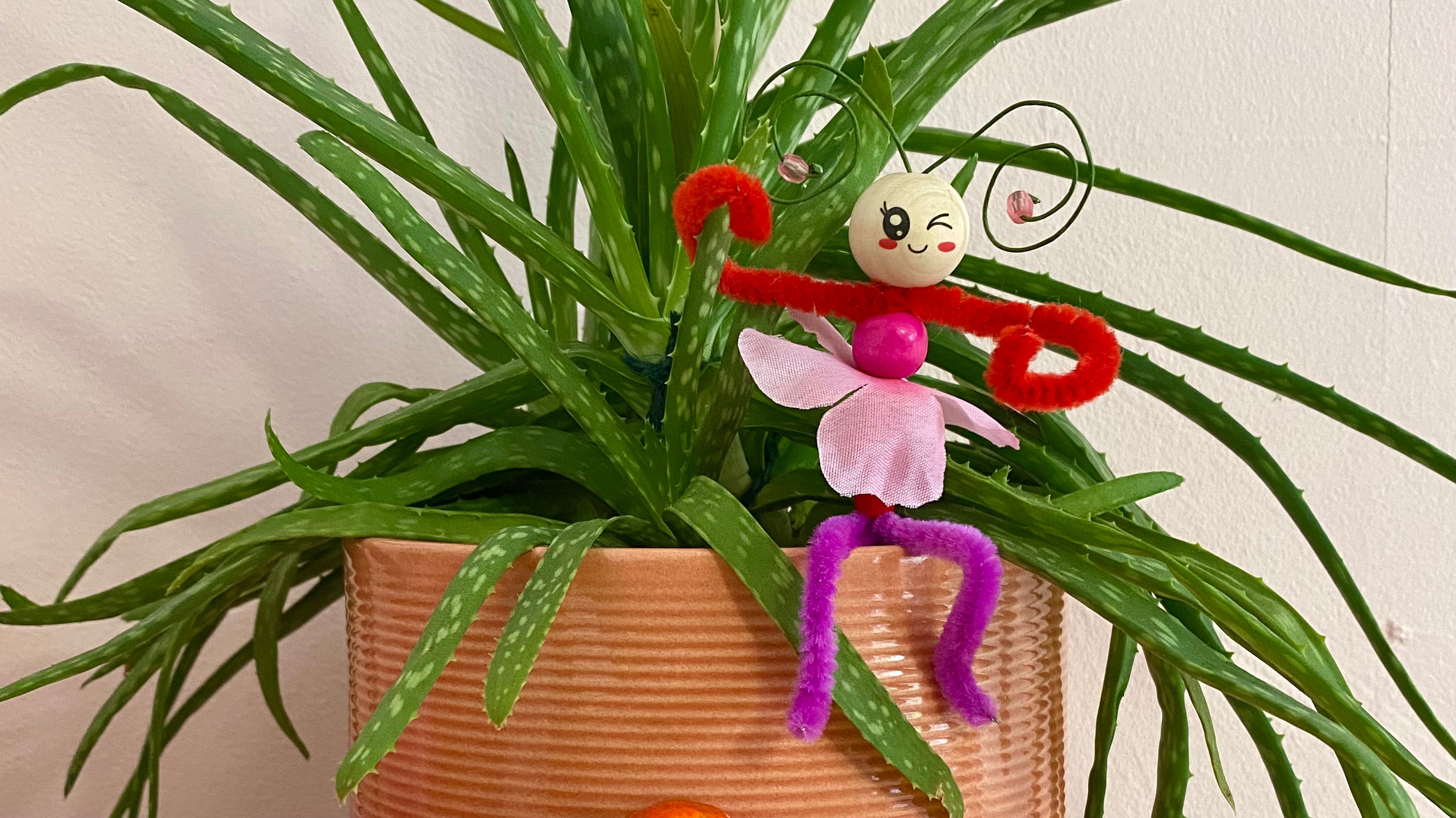 A fairy made from colourful pipecleaners and beads sits on the edge of an orange plant pot with one arm holding onto the plant. The face is winking.