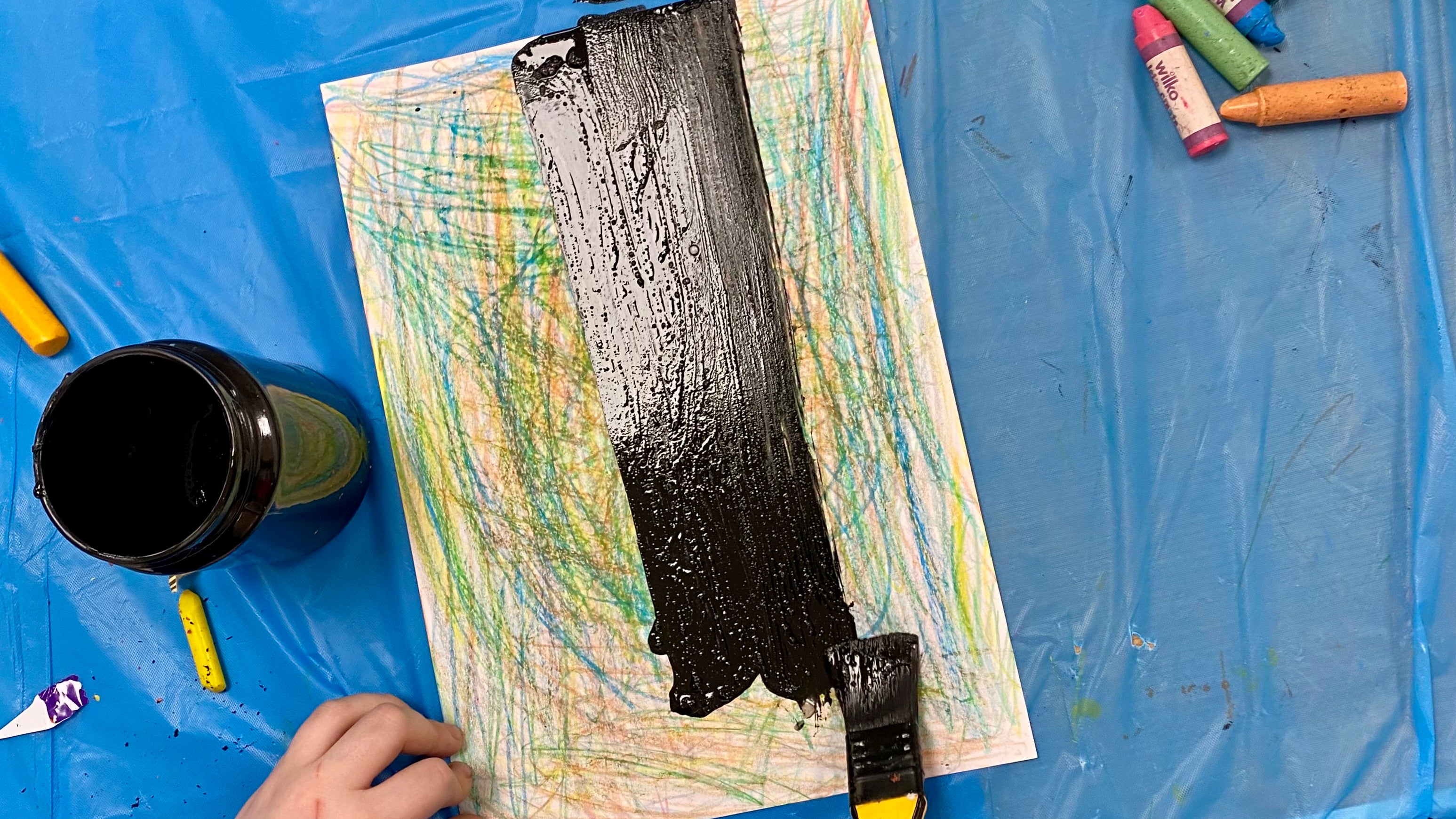 A hand holding a thick paintbrush loaded with black paint, painting black over colourful scribbles of wax crayon drawn on white card