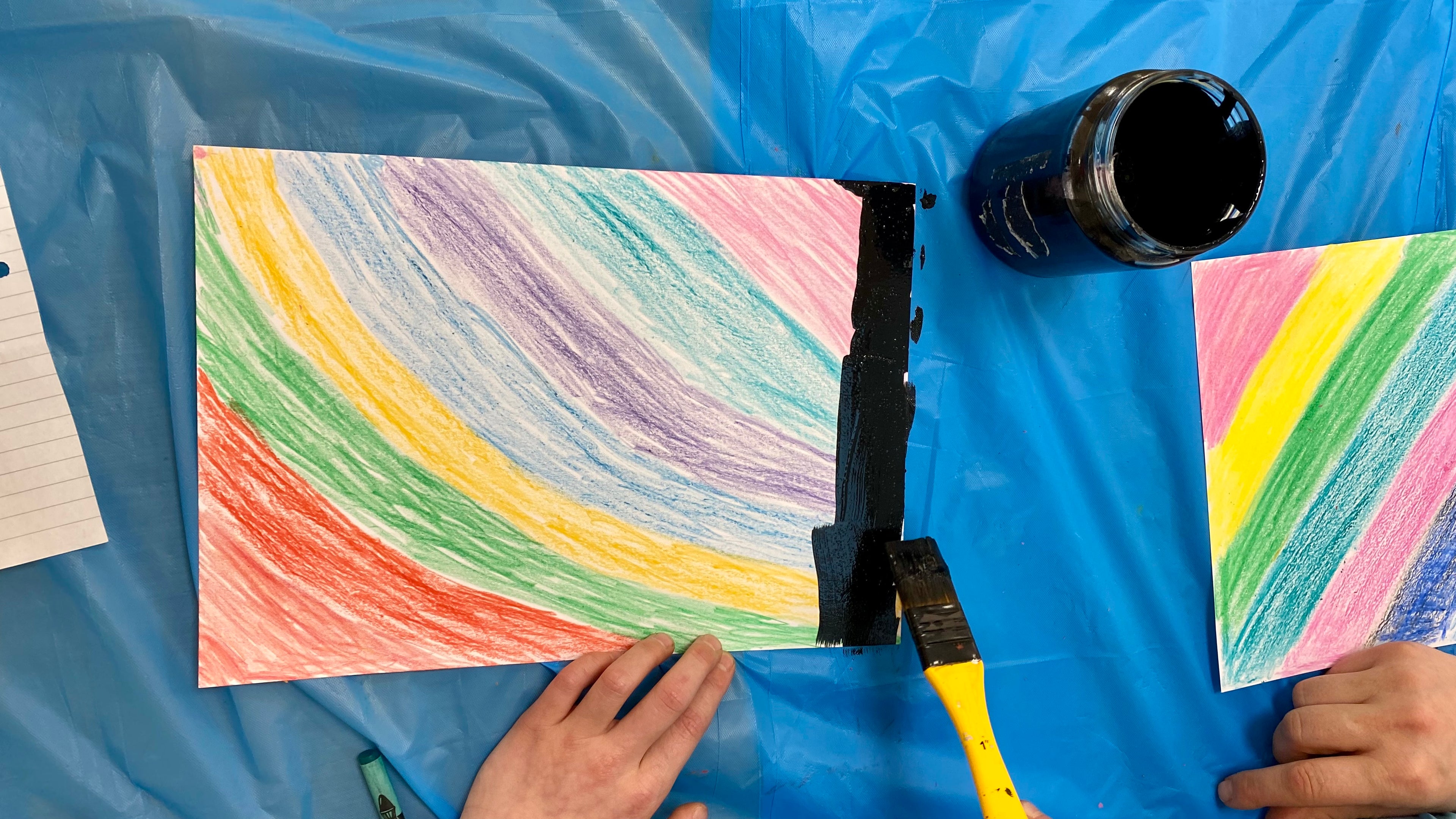 A hand holding a thick paintbrush loaded with black paint, painting black over colourful stripes of wax crayon drawn on white card