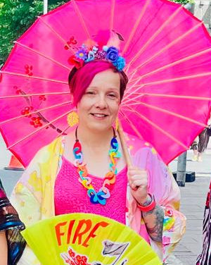 A women posing in vibrant, colorful festival attire on a city street, holding a decorative pink parasol. She wears a colourful floral head crown with a my little pony figure on.