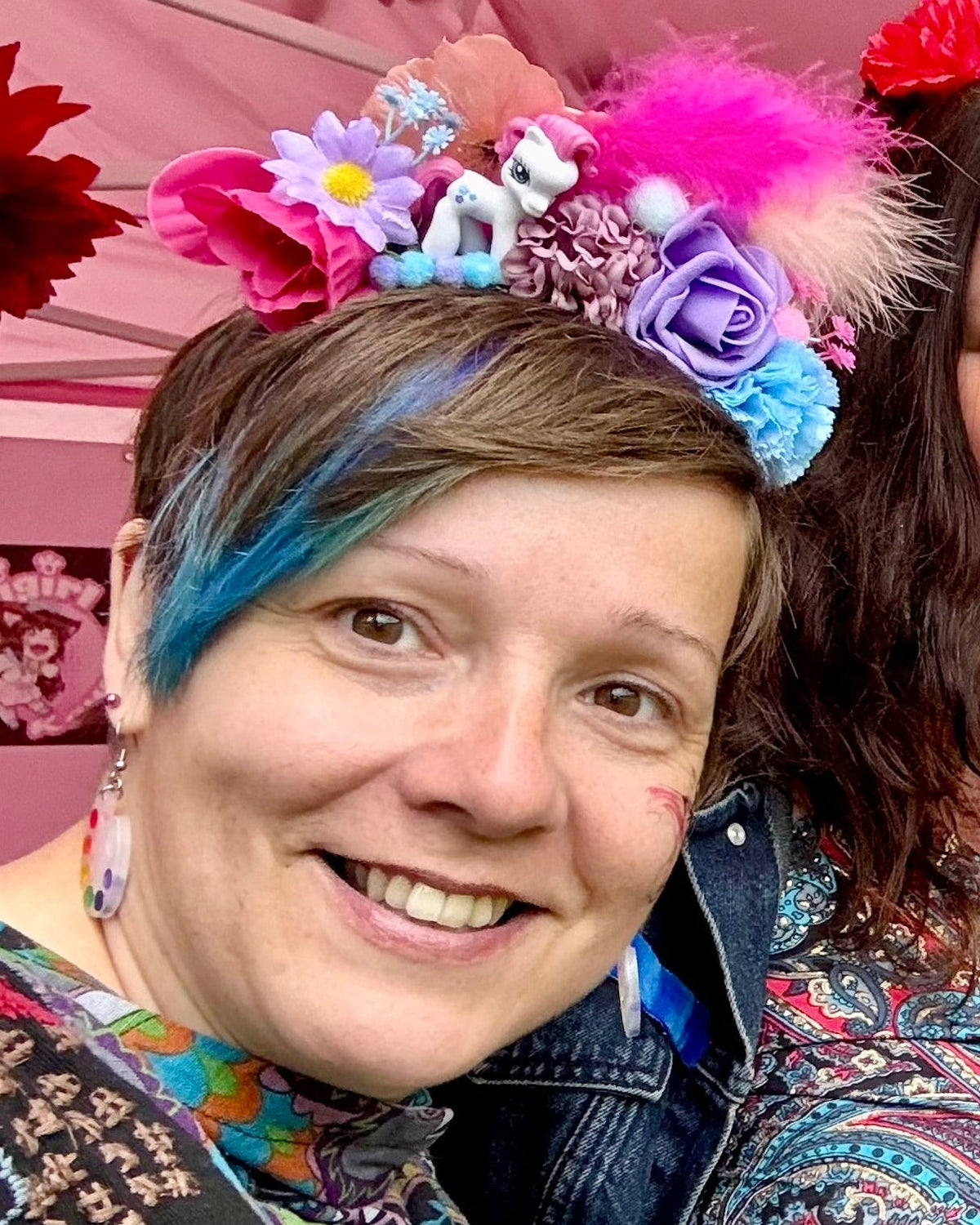 Two people smiling under a pink canopy at an outdoor event, each wearing a colourful floral headpiece. The closest to the camera is wearing a pink and purple flower and feather crown with a my little pony on.