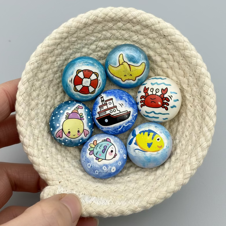 a small white rope bowl containing 7 colourful mini pebbles with various ocean themed designs on. A stingray, a life buoy, a rainbow lobster, a ship, a crab, a rainbow fish and a yellow fish.