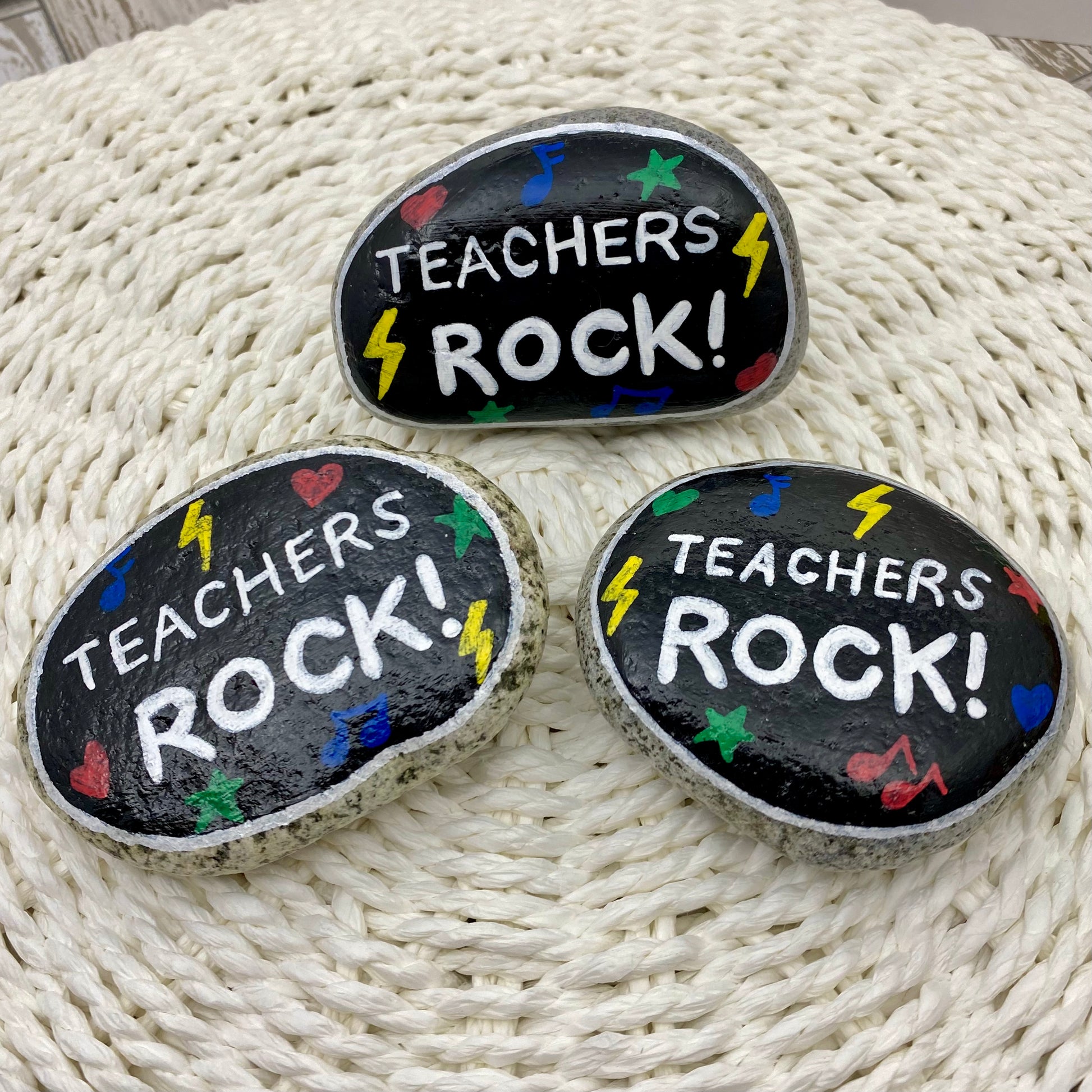 3 stones painted black with rock and roll symbols painted in bright colours and the words "Teachers Rock!" in white