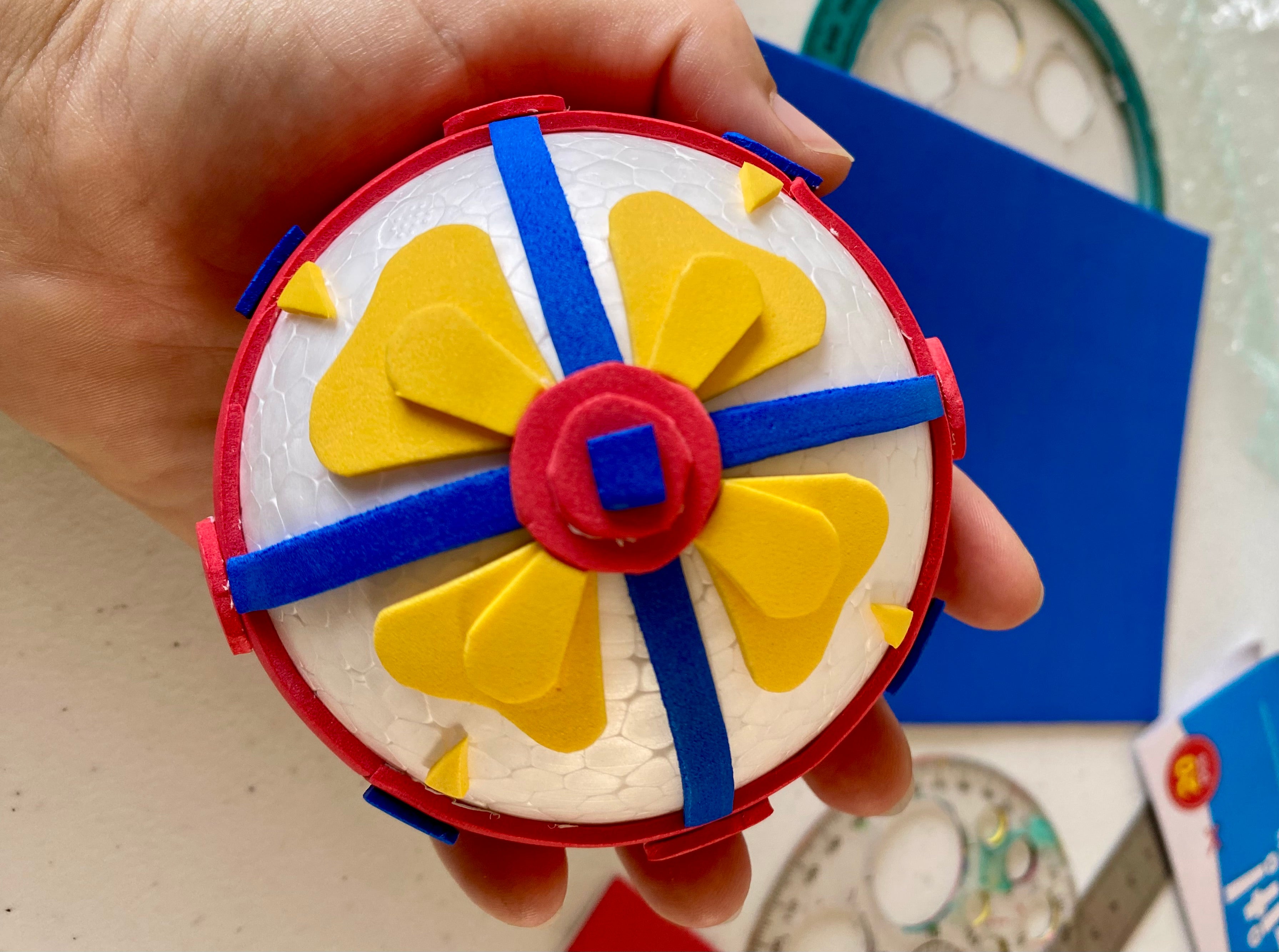 A white polystyrene half sphere covered with cut out pieces of red, yellow and blue coloured craft foam arranged to make 3D shapes to resemble one of the ceiling bosses from inside Exeter Cathedral