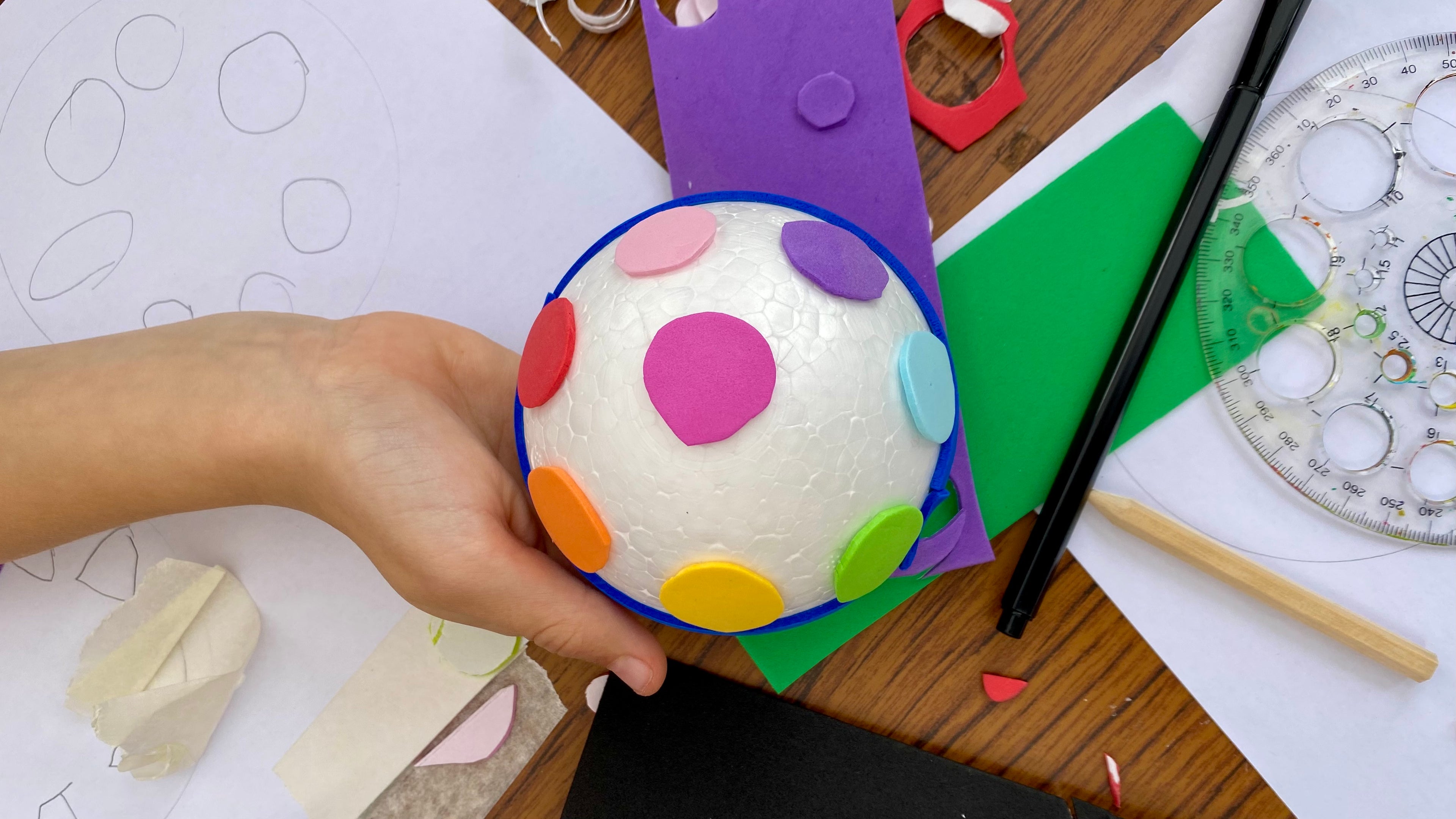 A white polystyrene half sphere covered with circles of coloured craft foam stuck on, being held above a table covered in crafting materials.