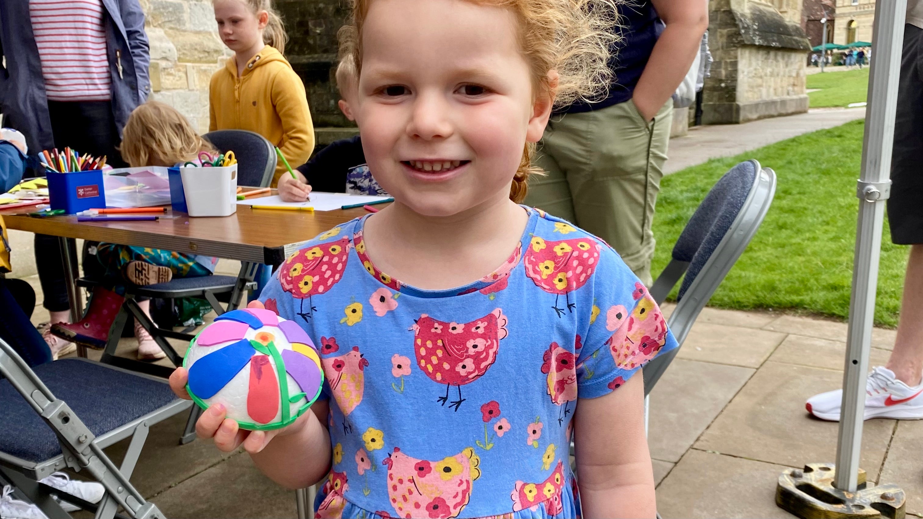 A white polystyrene half sphere covered with cut out pieces of coloured craft foam arranged to make a flower being held by a girl. In the background a busy craft table with other children making their creations in front of the cathedral
