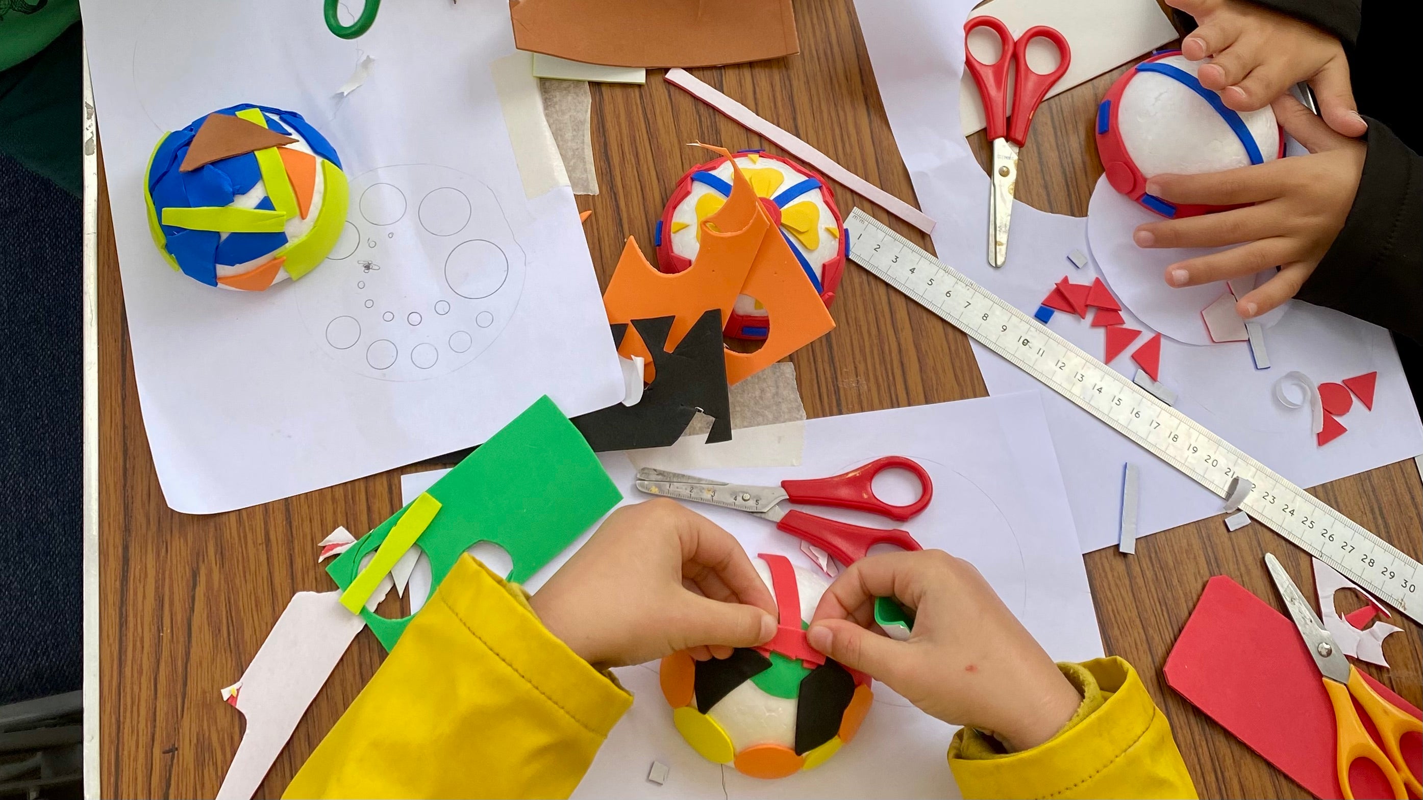 A table with four white polystyrene half spheres being covered with shapes of coloured craft foam, surrounded by crafting materials.