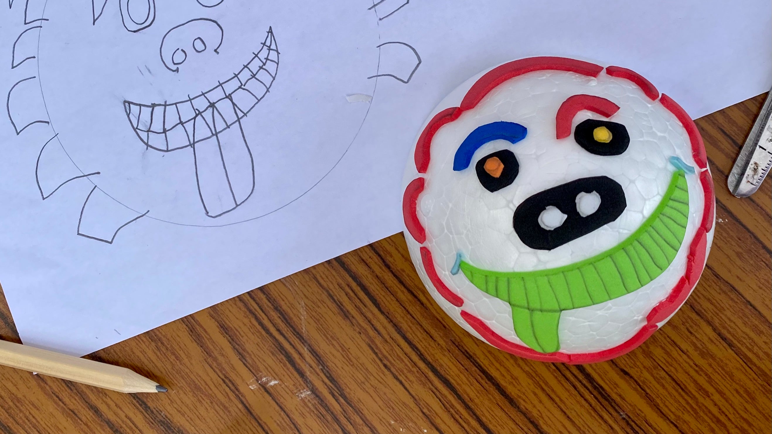 A white polystyrene half sphere covered with cut out pieces of coloured craft foam arranged to make a gargoyle face, on a table next to a worksheet with a drawing of the design.