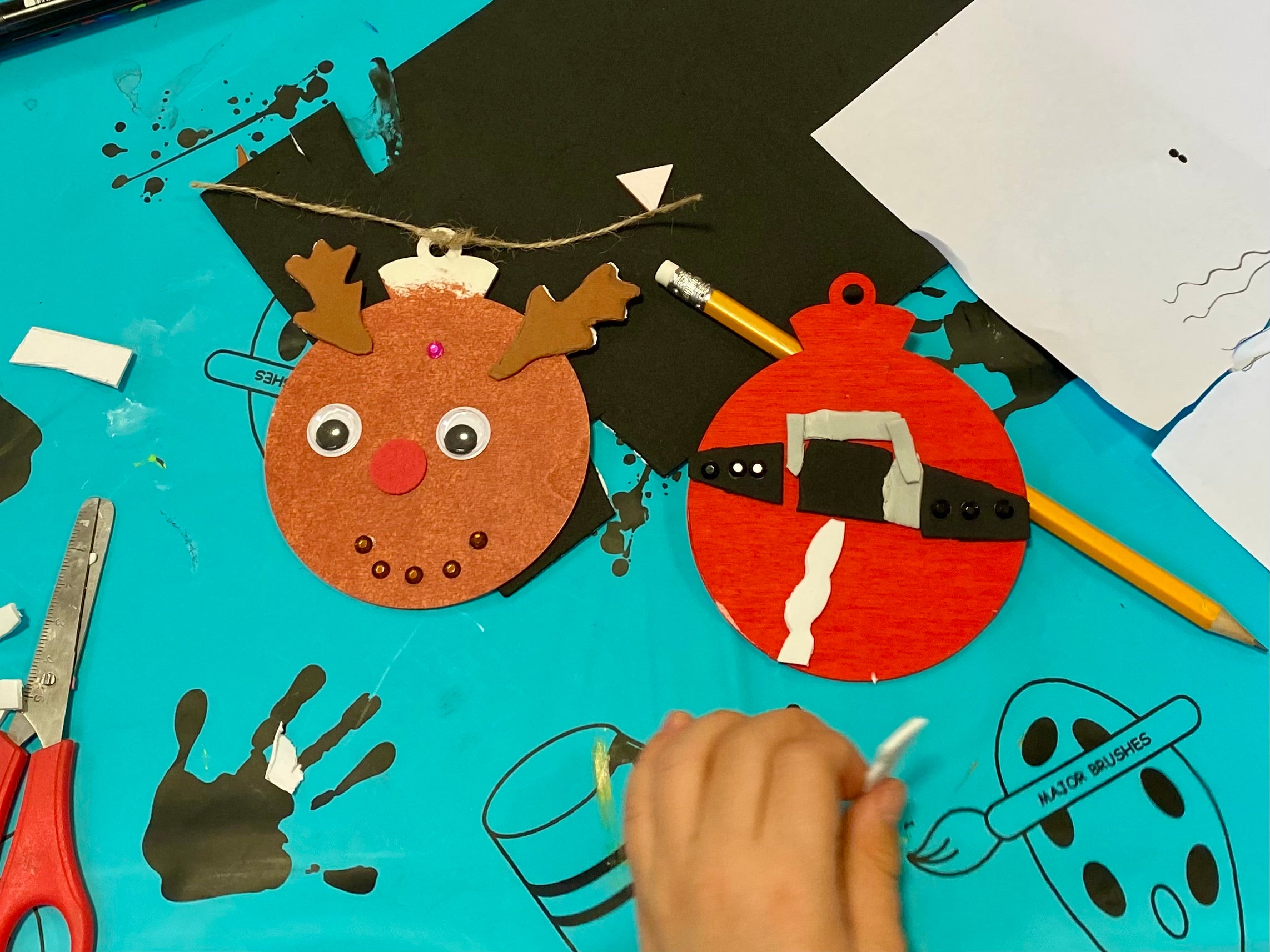 2 painted baubles on a crafting table. One is a reaindeer with antlers, googley eyes and a red nose, and the other is a santa themed red bauble with white trim and a black and gold belt around.
