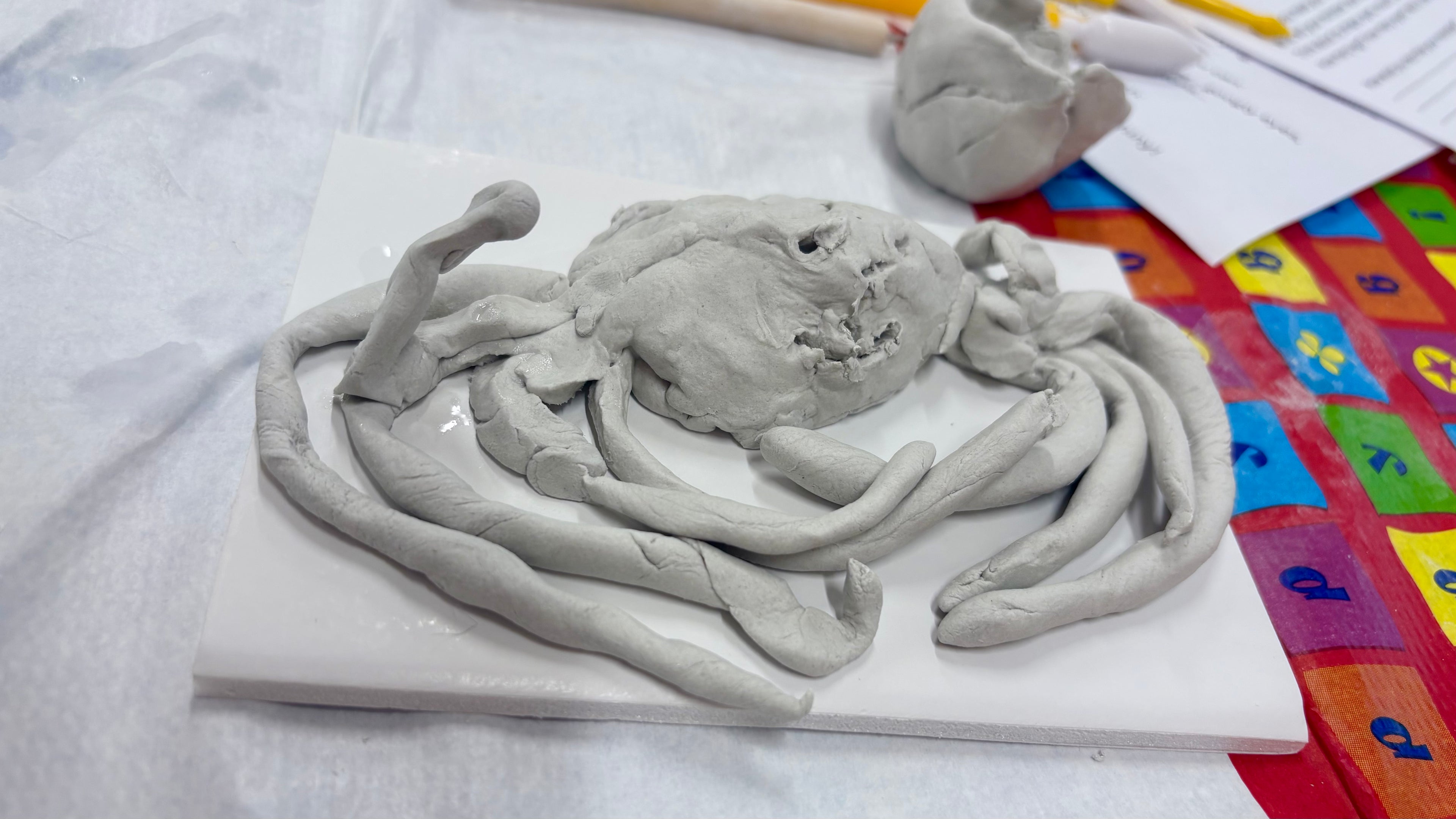 Clay sculpture of a creature with many legs on a white surface with colorful patterned tablecoth underneath