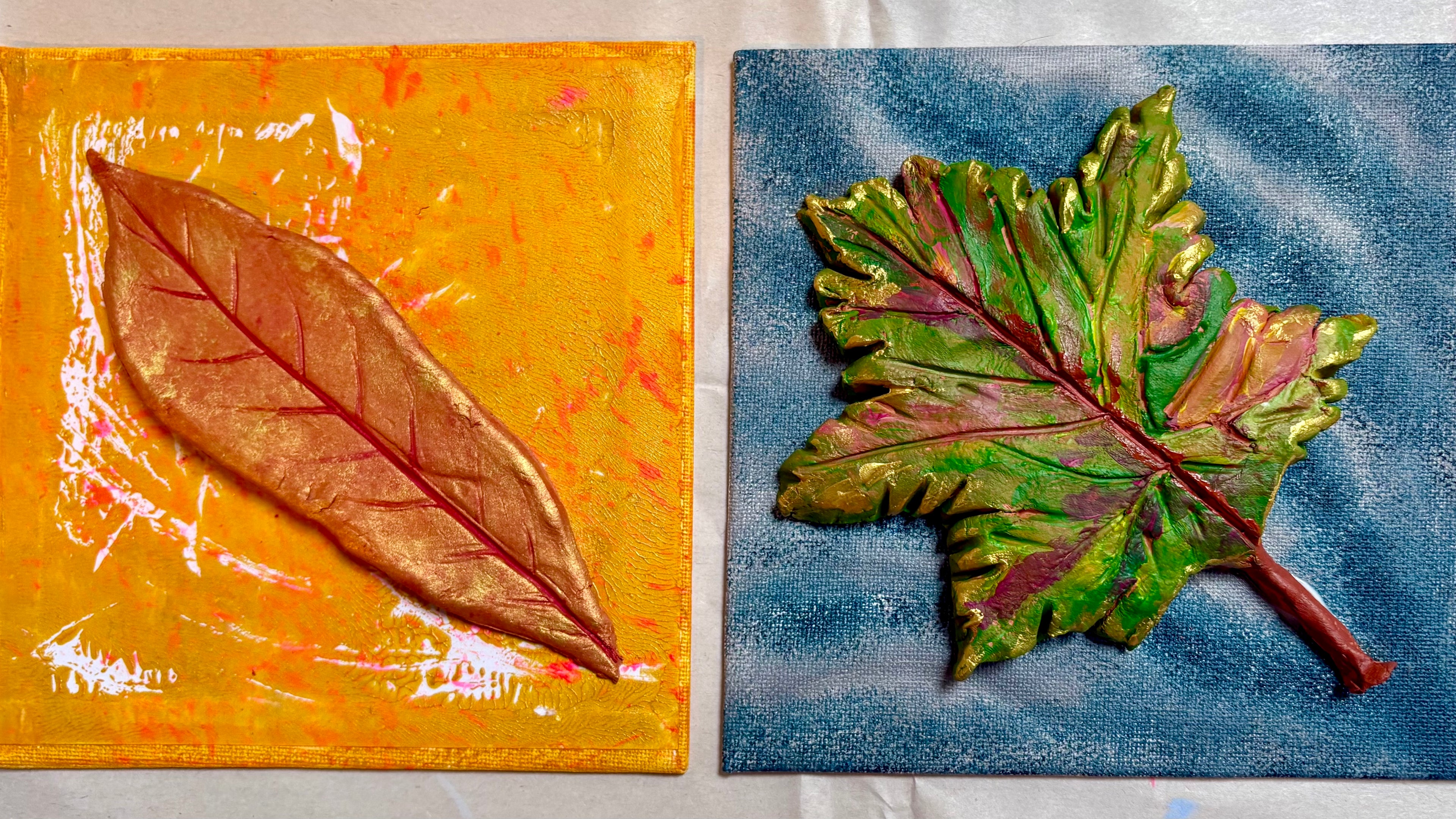 Two square painted canvases side by side, each featuring a large clay leaf motif. Painted in bright colours with gold accents.