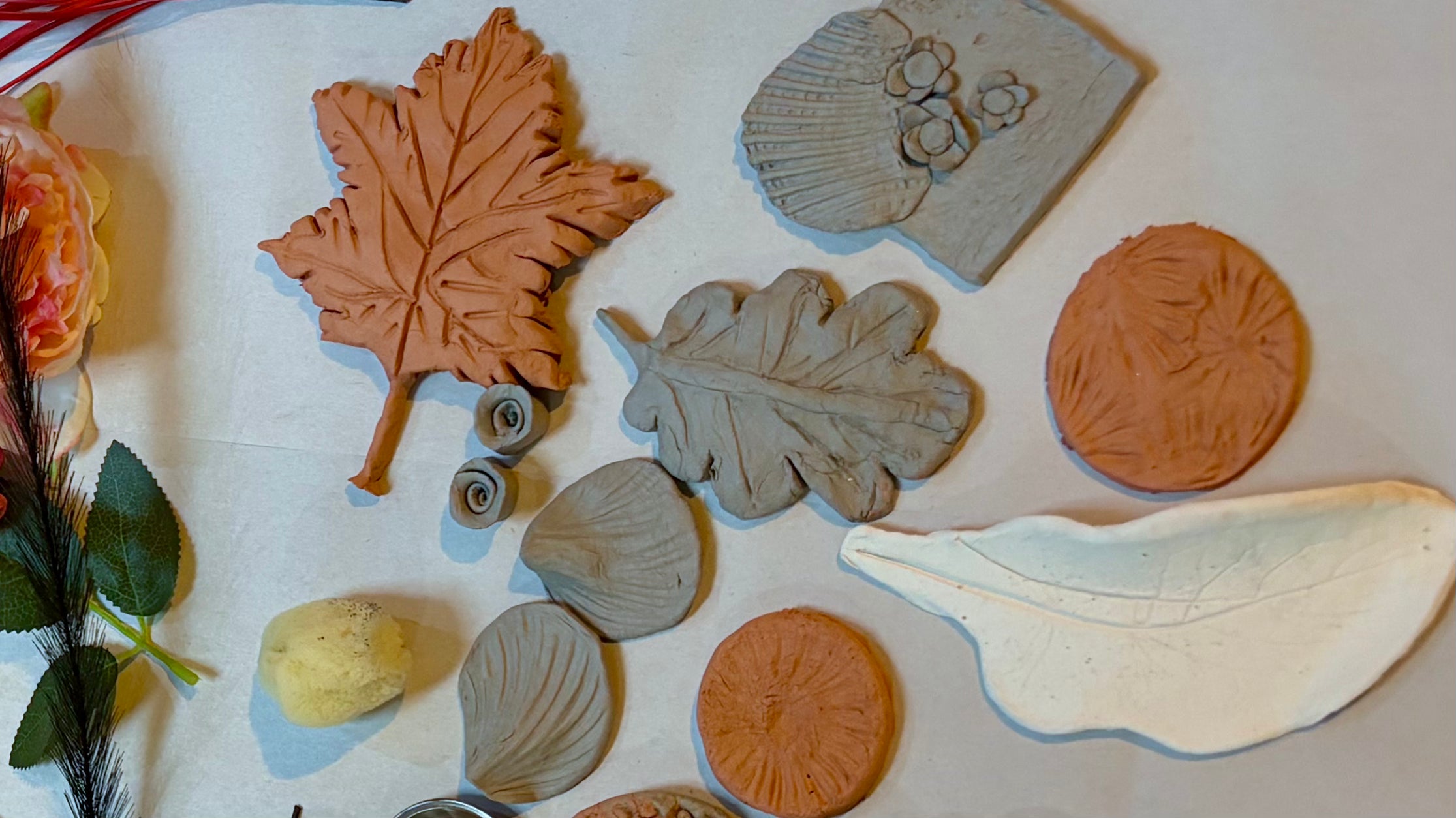 A flat-lay display of various clay shapes including leaves, shell, and circular botanical designs in terracotta, stone and white air dry clay, arranged on a workbench.
