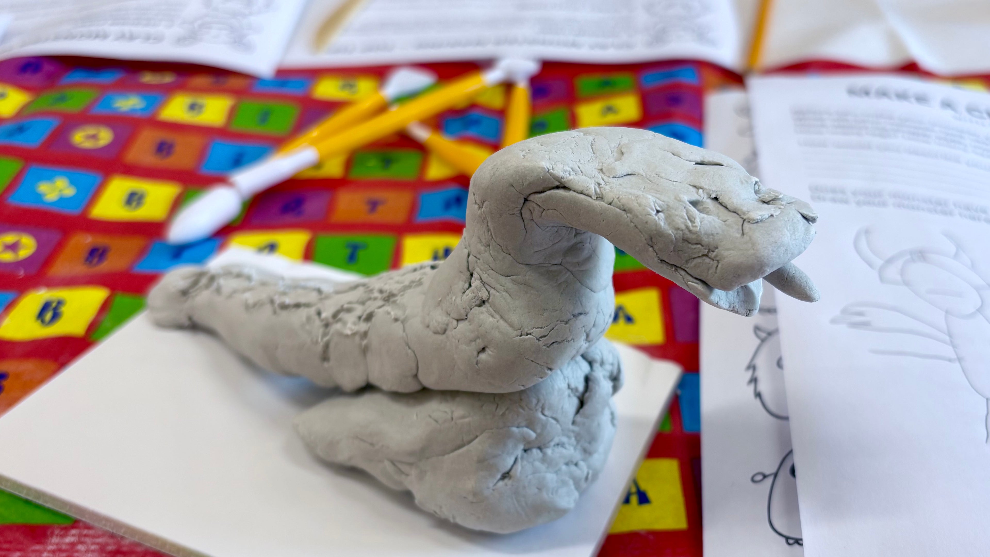 Clay sculpture of a snake on a piece of paper with colorful tablecloth and clay tools in the background.