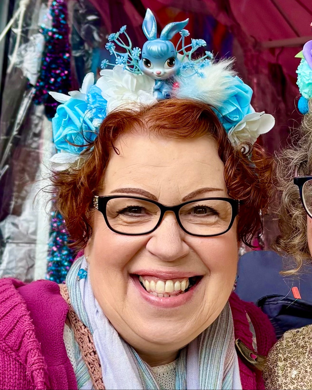 A smiling women standing outdoors at a festival under a pink canopy with sequined clothes behind her. She wears a blue floral headpiece featuring a toy rabbit.
