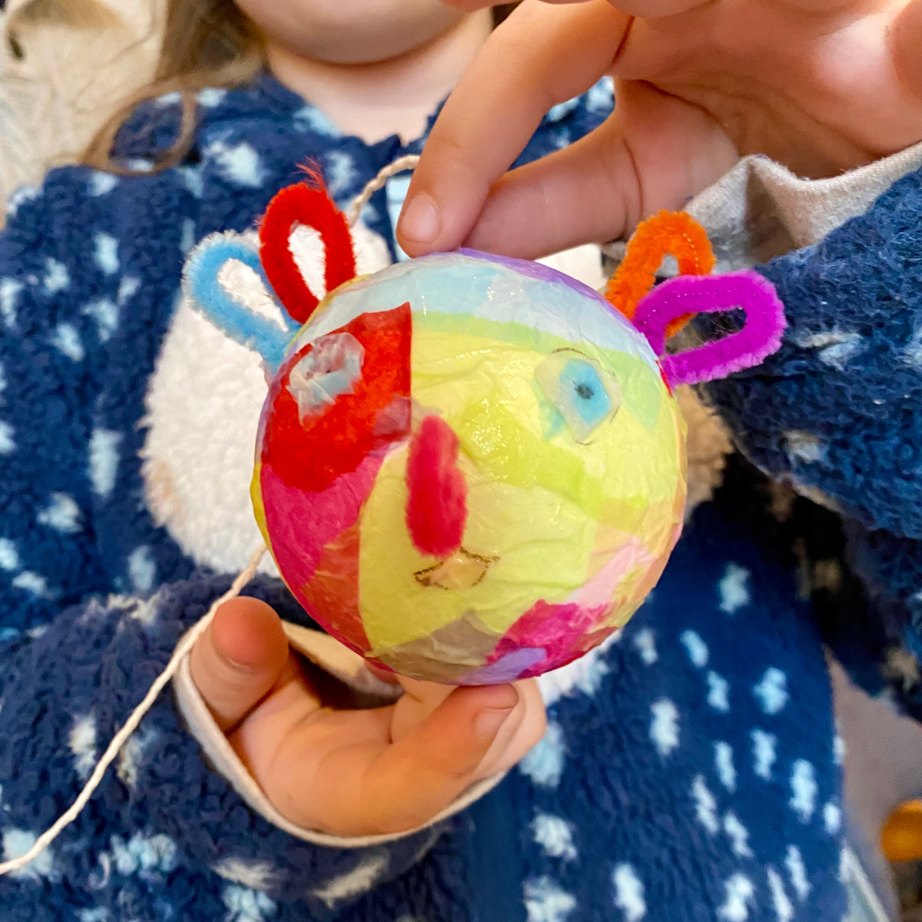 A paper mache planet creation being held by a child. The planet looks like a face and is covered with brightly coloured tissue paper and has pipe cleaners attached for ears, eyes and a nose.