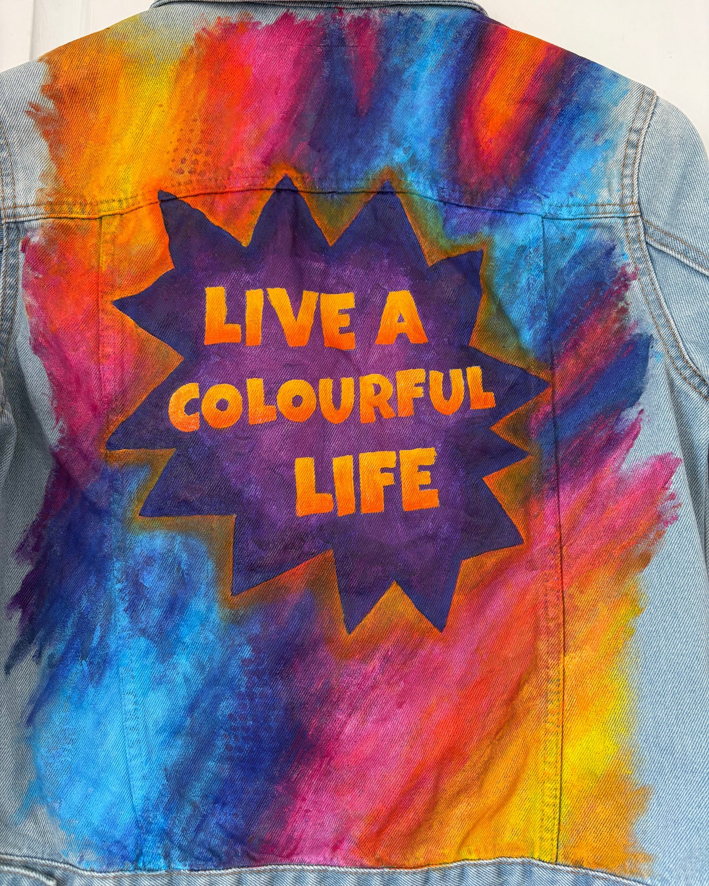 Close up of painted panel on Denim jacket. Colourful grafitti style design with 'Live a Colourful Life' text painted in a purple starburst