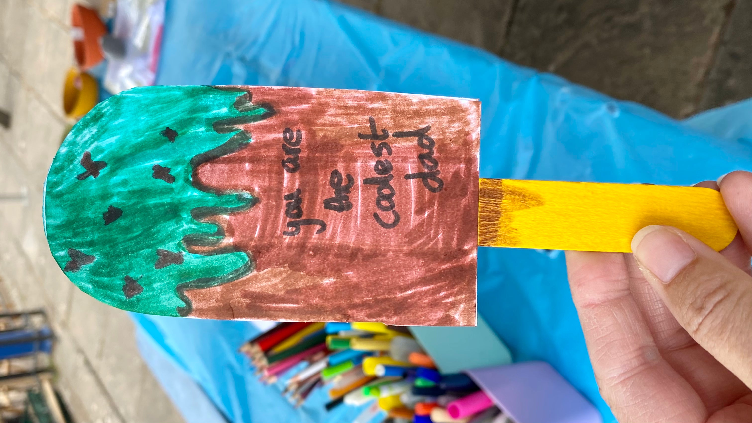 Ice Lolly card on a yellow wooden stick being held in front of an art table with colouring supplies. The cards is brown and green to represent mint chocolate chip and has the writing: You are the Coolest Dad