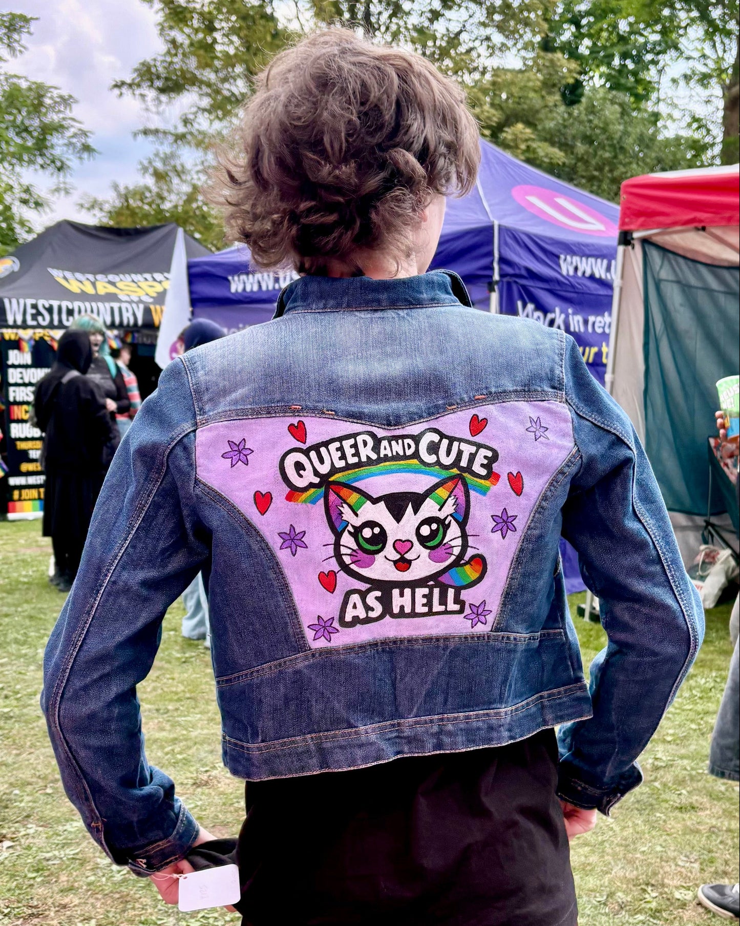 A person standing outdoors at a festival, viewed from behind, wearing a dark denim jacket with a large colourful painted graphic on the back that reads 'QUEER AND CUTE AS HELL' with a cartoon cat. Other people, tents, and trees are visible in the background.