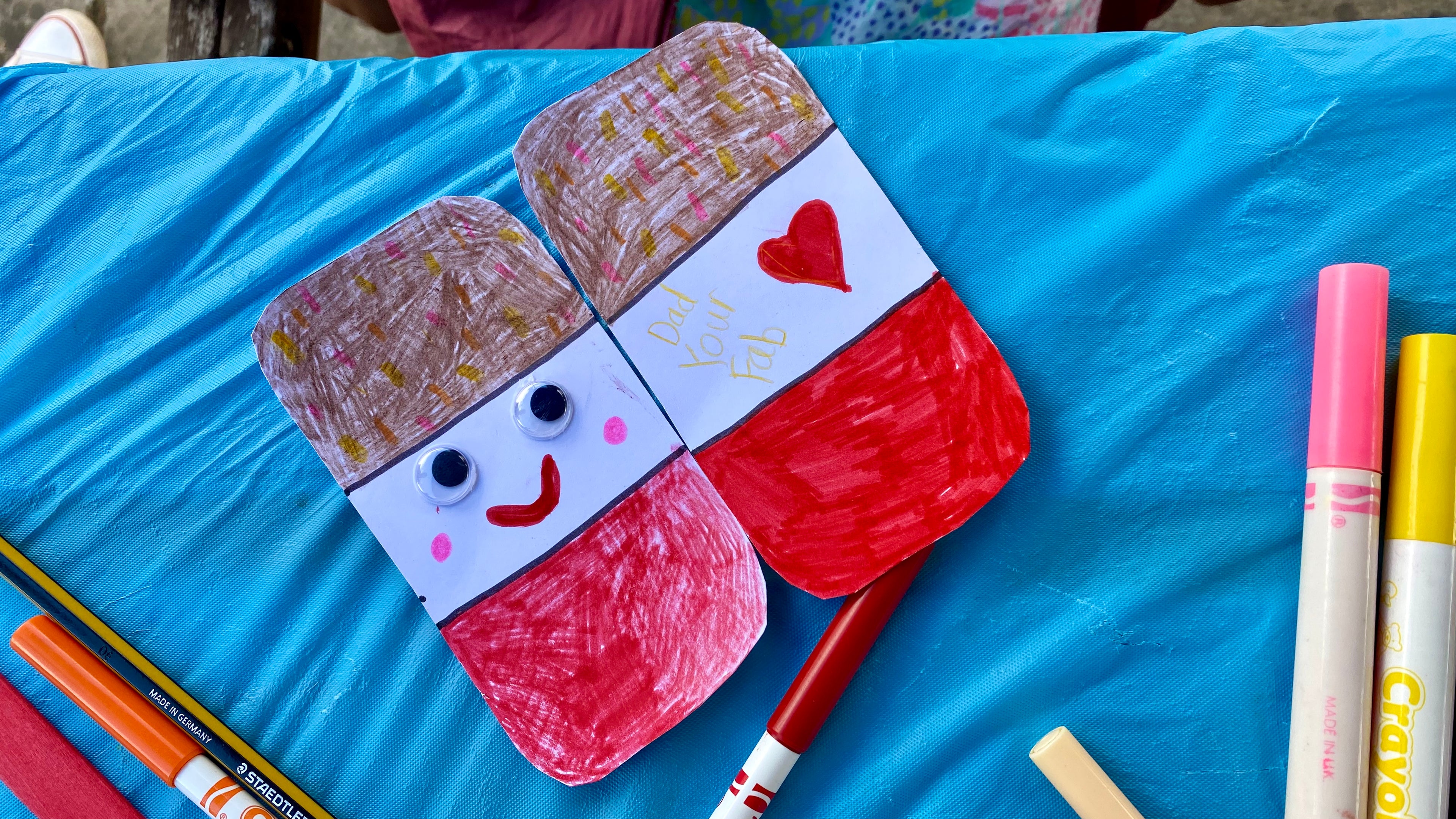 2 sides of a card in the process of being coloured in. The card looks like a Fab ice lolly with a smiley face and heart on it, there are pens and pencils also in the shot. 