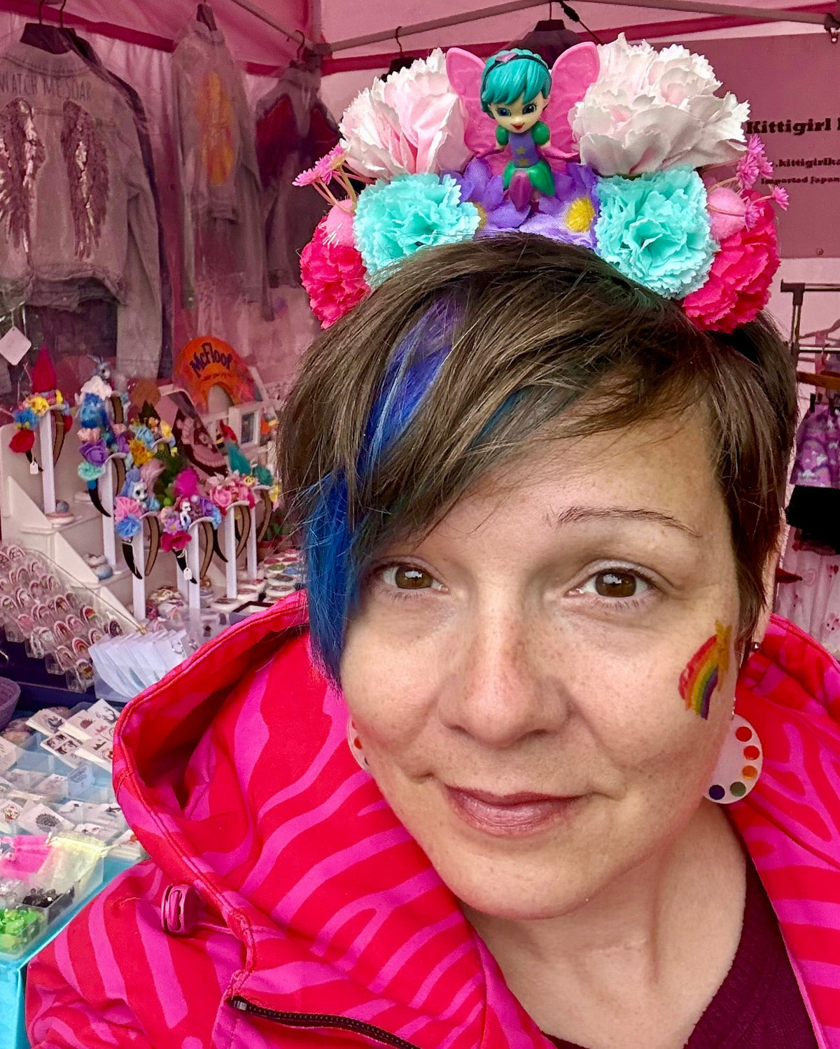 A woman at a market stall wearing a colourful floral headband with toy fairy figure, dressed in a pink patterned jacket, with more flower crowns and painted denim jackets in a pink gazebo in the background.
