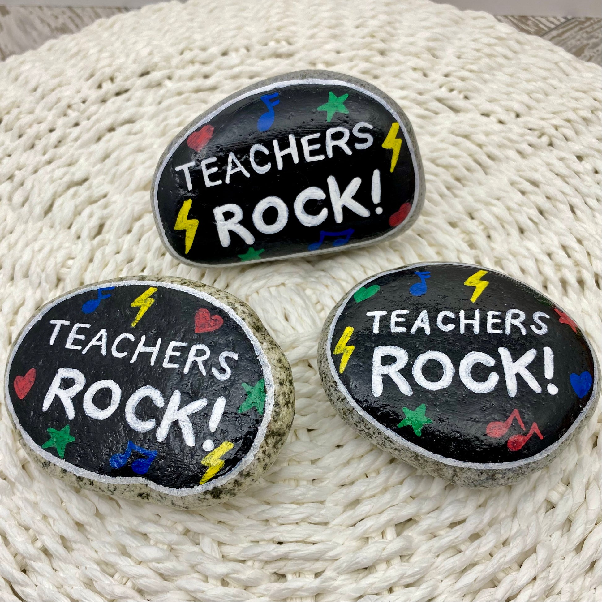3 stones painted black with rock and roll symbols painted in bright colours and the words "Teachers Rock!" in white