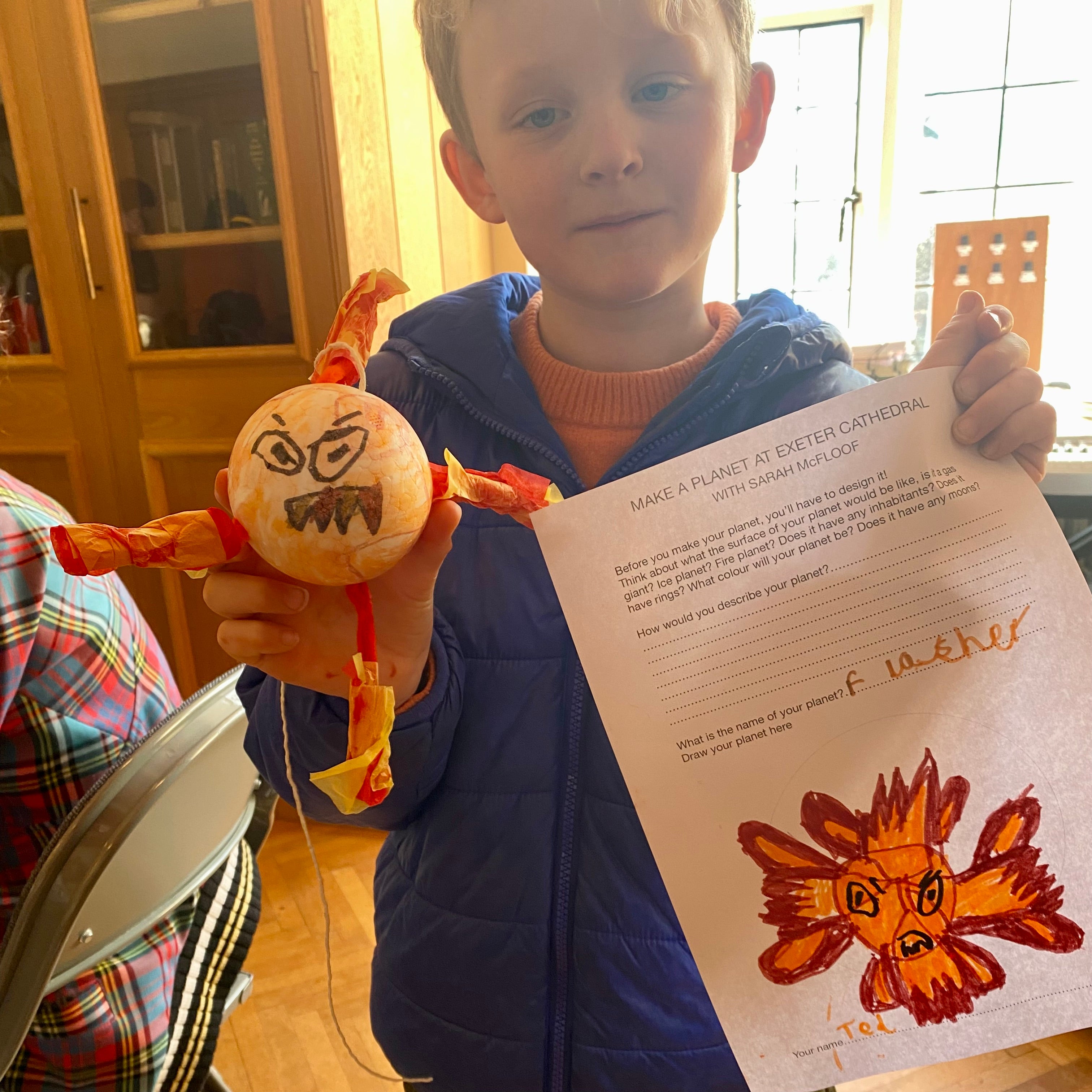 A boy holding up his planet creation and planning worksheet. The planet has fiery paper mache plumes coming out from it and an angry face drawn on the surface.