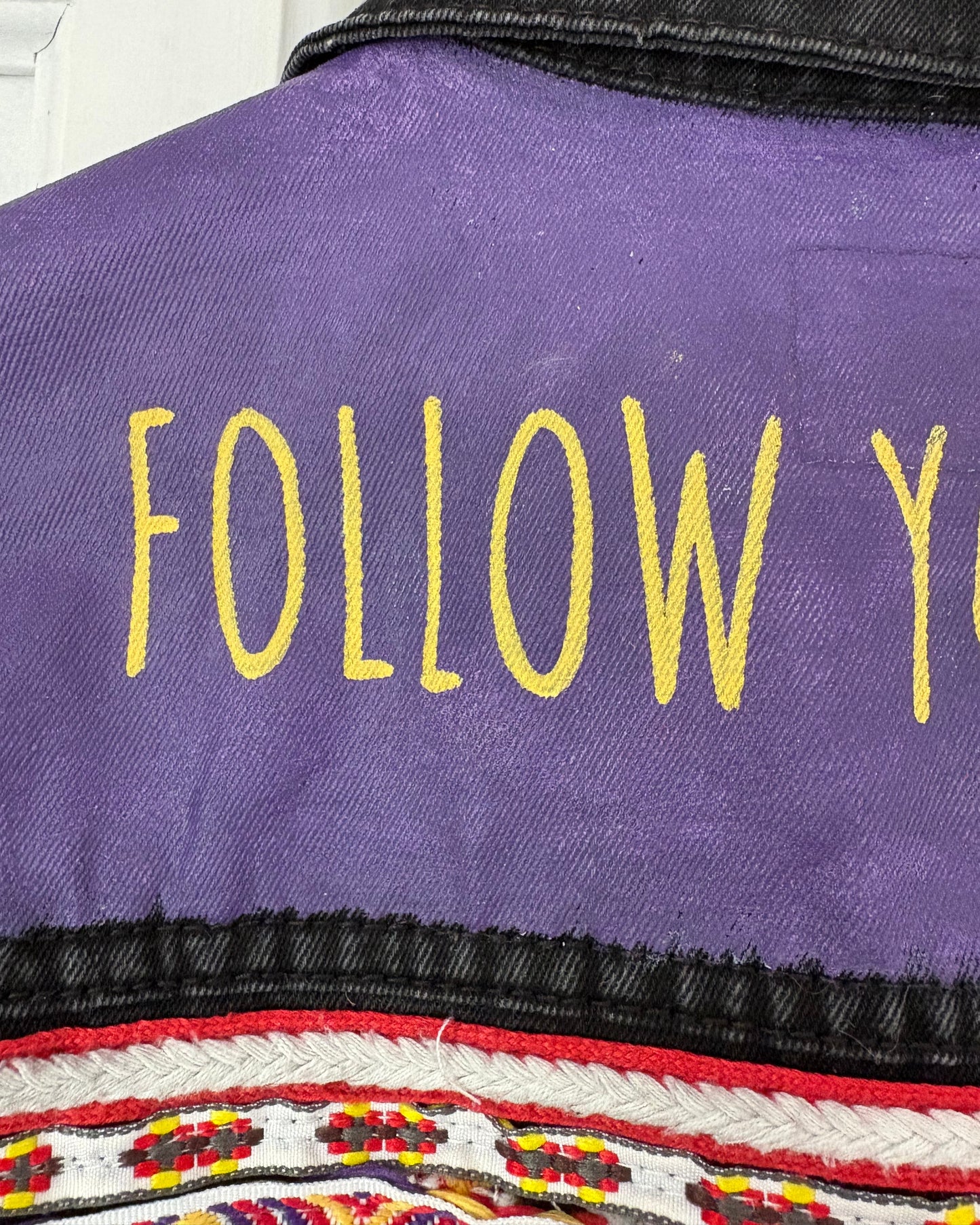 Purple painted panel on a black denim jacket with yellow text 'FOLLOW' and a colourful patterned embroidered ribbon underneath