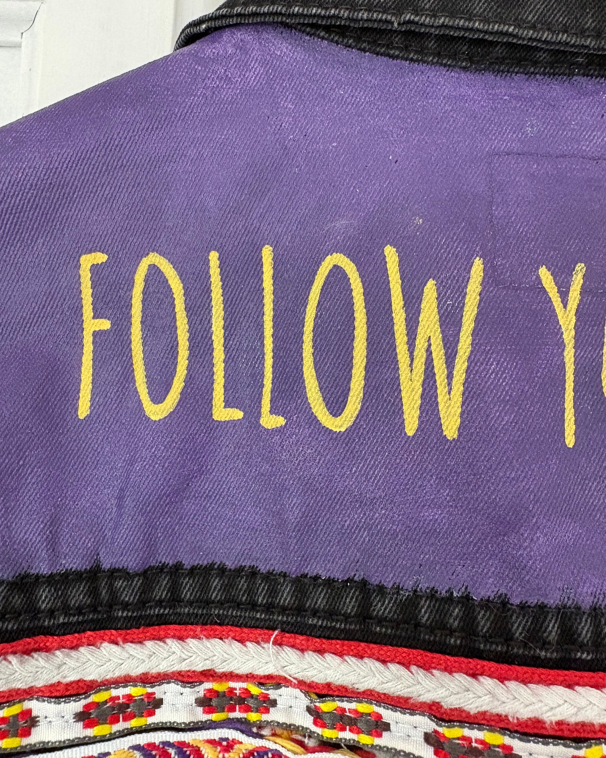 Purple painted panel on a black denim jacket with yellow text 'FOLLOW' and a colourful patterned embroidered ribbon underneath