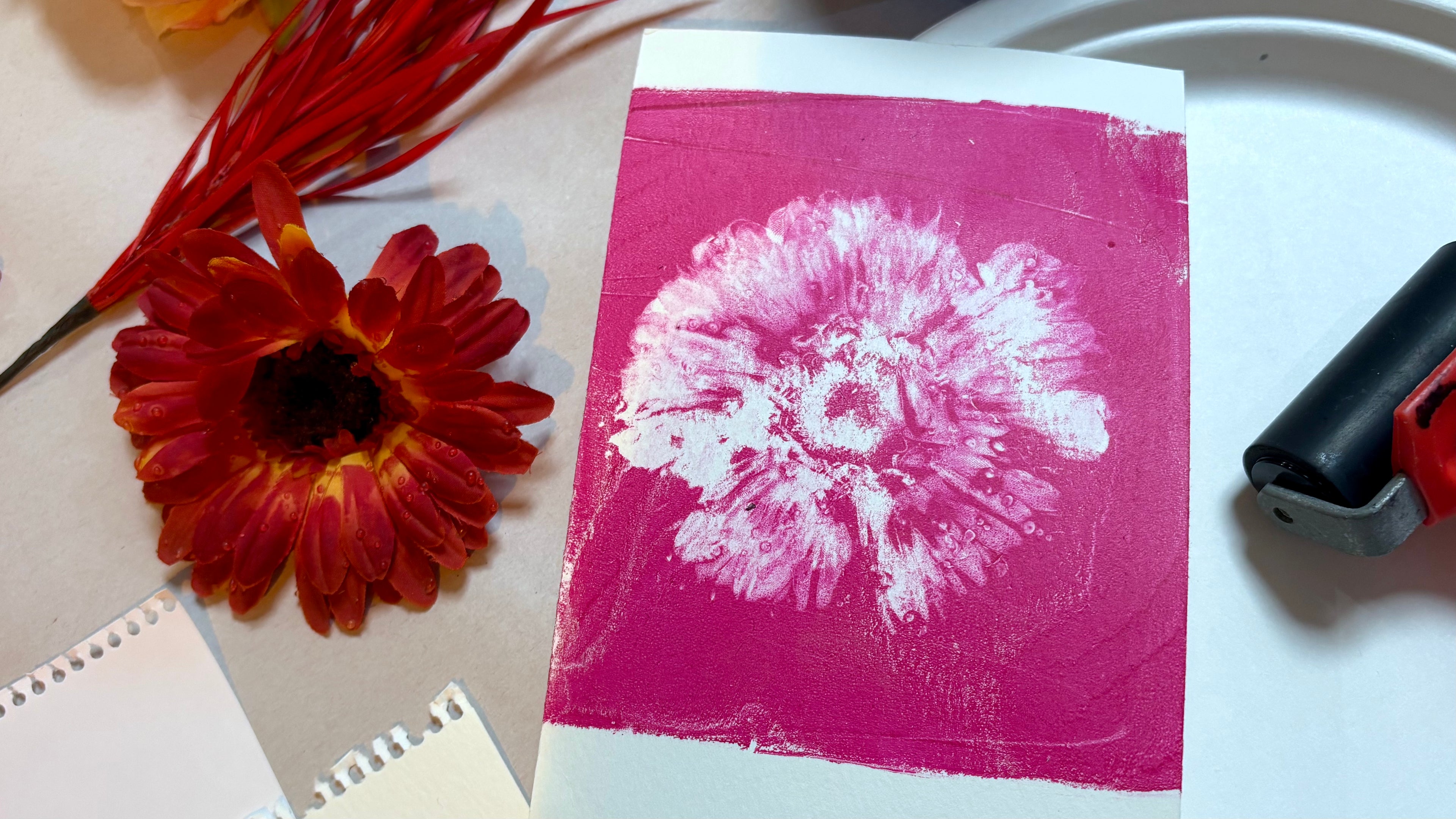Pink print of a large flowerhead next to the painted flower used to make the print. Also a paint roller off to the side.