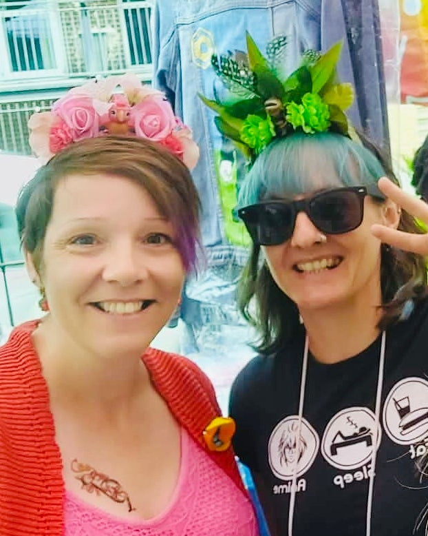 Two women smiling for a selfie at a market, with colorful clothing displayed on mannequins and racks behind them. One wears a green feather, leaf and flower crown with a fairy on.